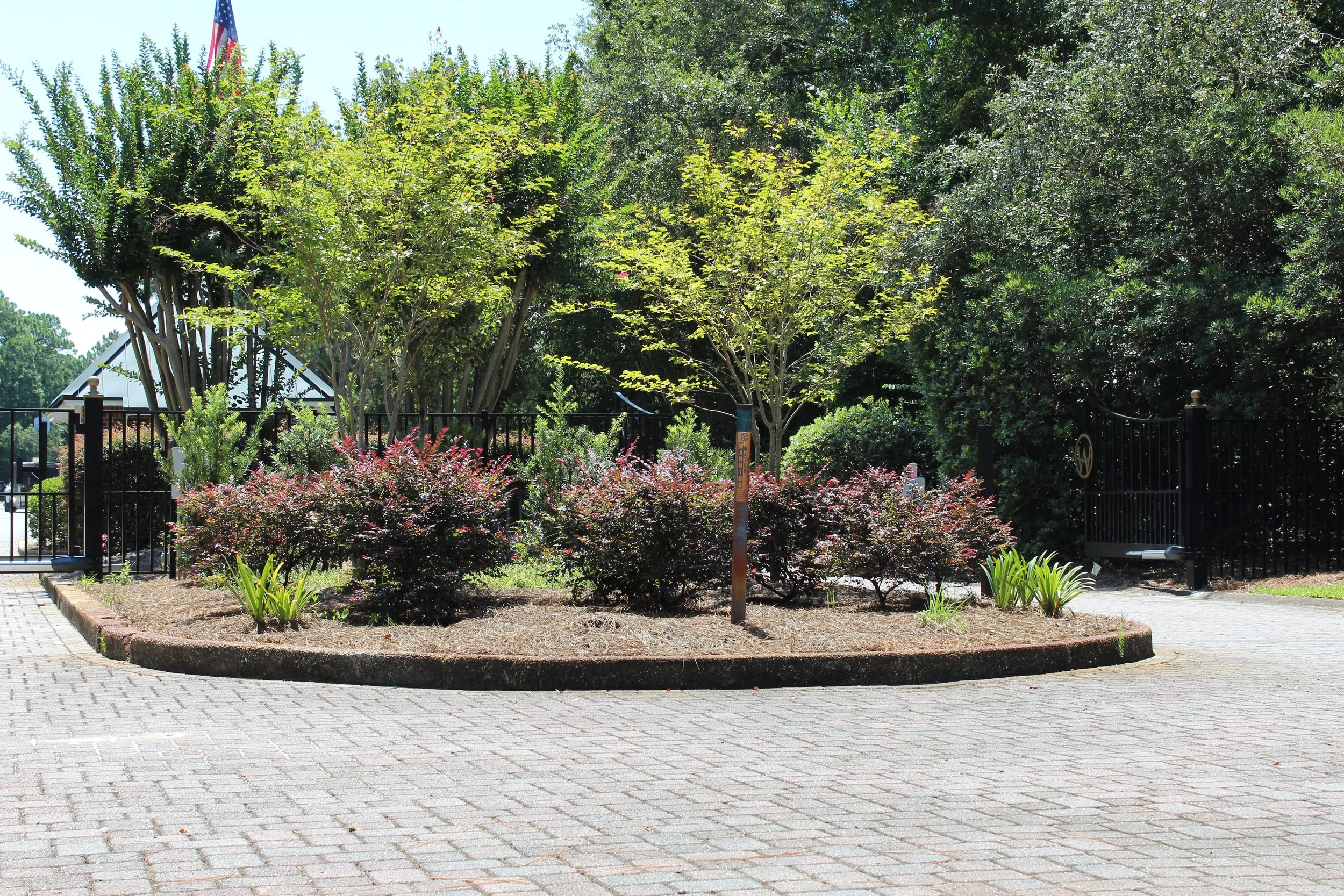 View of a landscaped garden area with various green trees and bushes, a brick-paved walkway in the foreground, a black metal fence surrounding the garden, and a small American flag visible in the background.