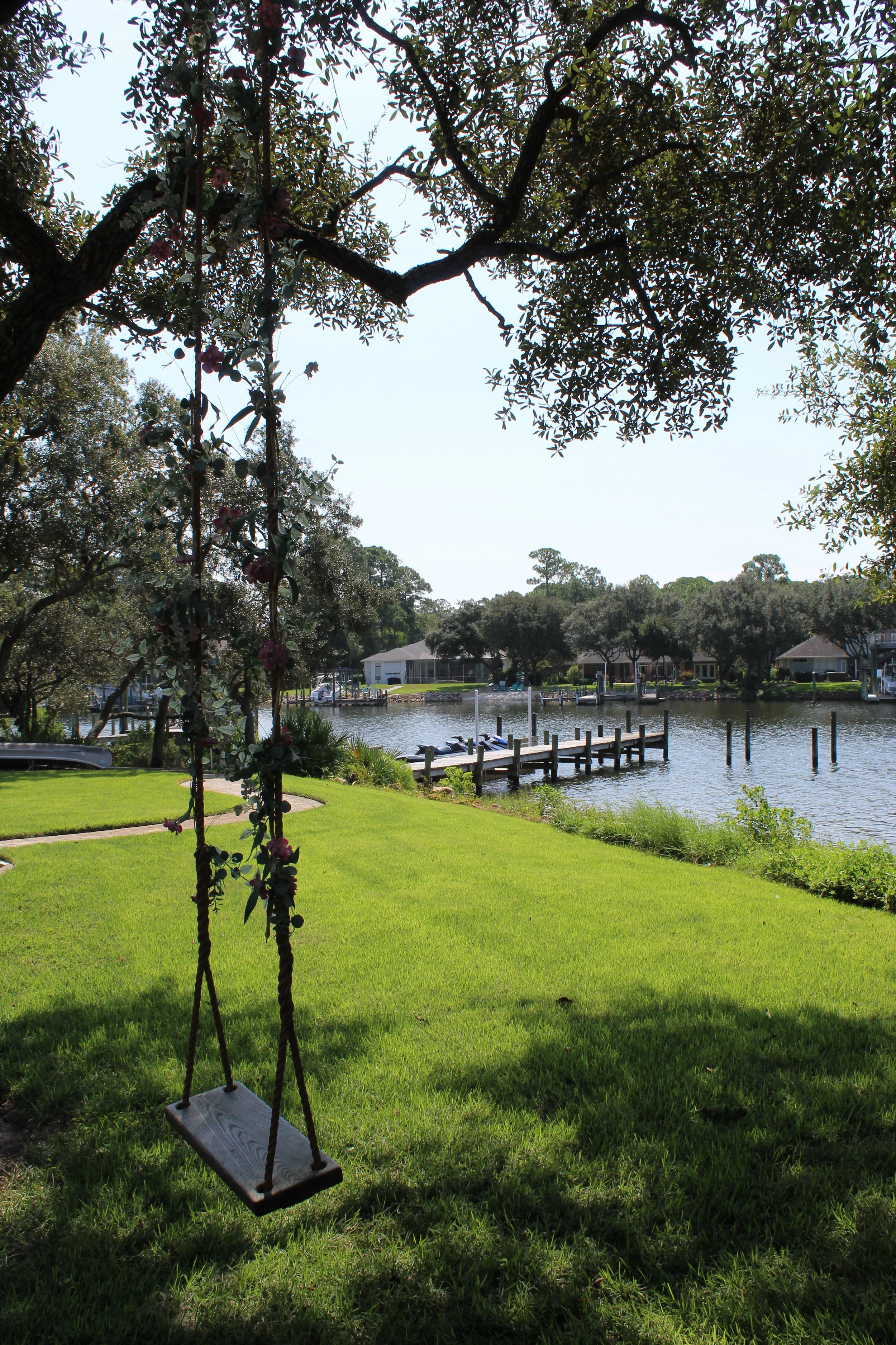 A backyard scene with a wooden swing hanging from a tree branch in the foreground. The yard has lush green grass and a body of water with wooden docks and boats, surrounded by houses and trees in the background.