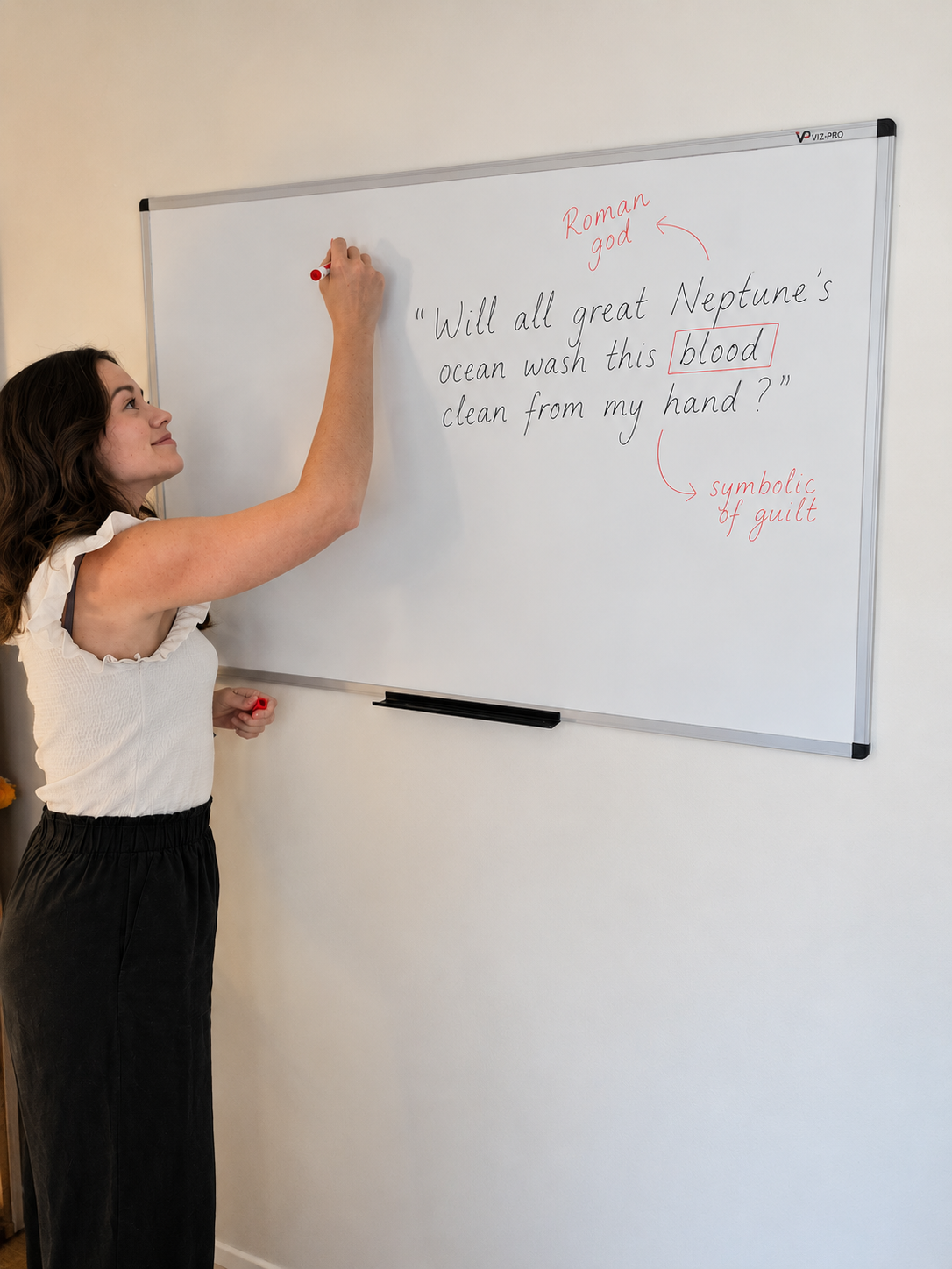 A woman writing on a whiteboard with a red marker. The whiteboard displays a quote from Shakespeare's 'Macbeth' about Neptune, with annotations in red indicating 'Roman god,' 'blood,' and 'symbolic of guilt.'