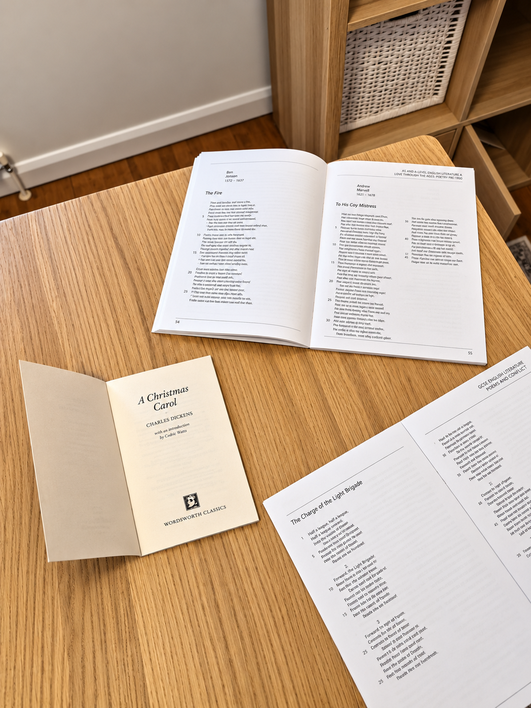 Open books on a wooden table, including a poetry collection, a Dickens novel, and a poetry pamphlet. The background features a wooden shelf and a corner of a room.