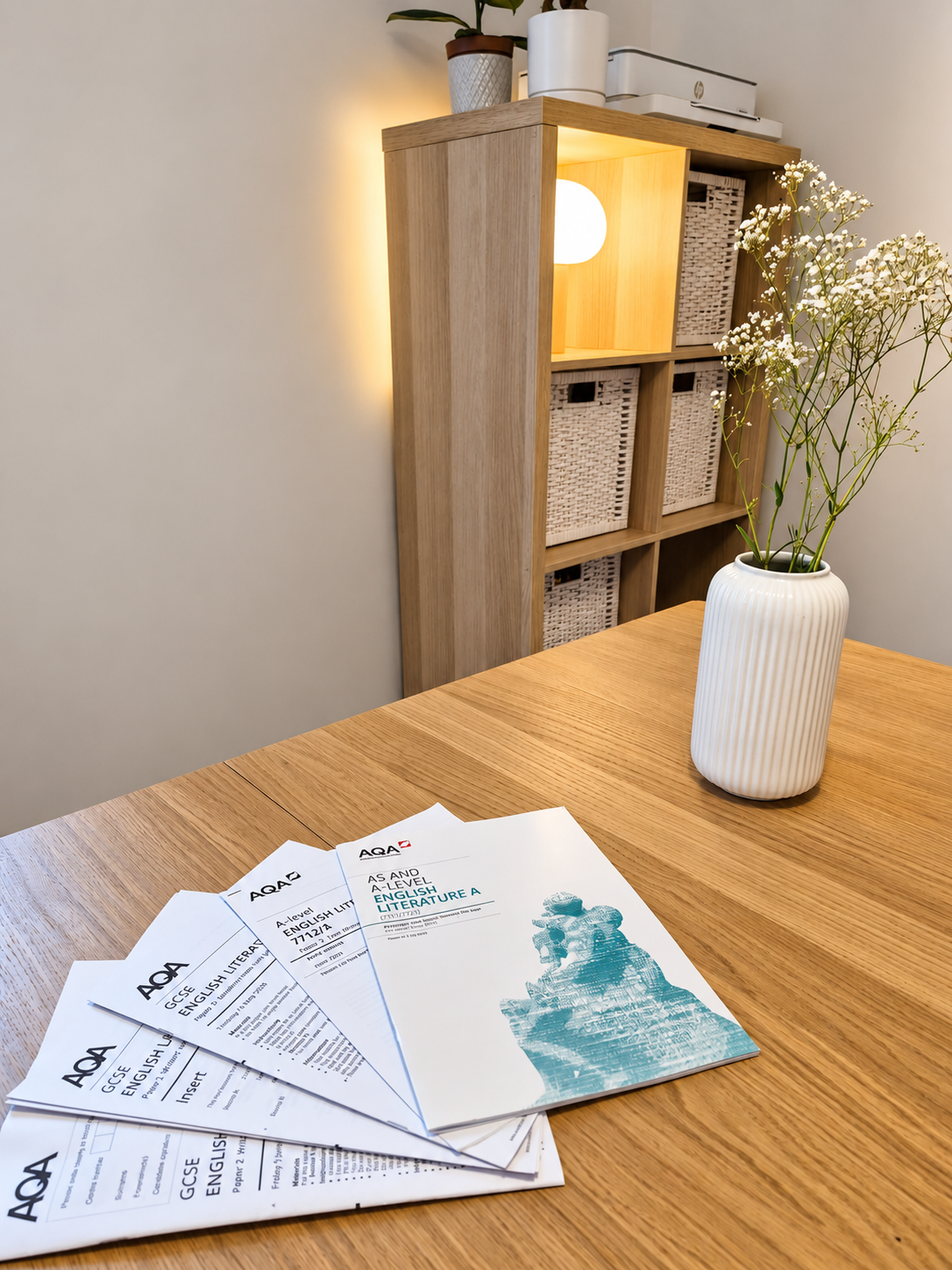 A wooden table with educational papers titled 'AQA GCSE English Literature A' spread out. There is a white vase with white flowers on the table. In the background, a wooden shelf with baskets, a lit lamp, and potted plants is visible.