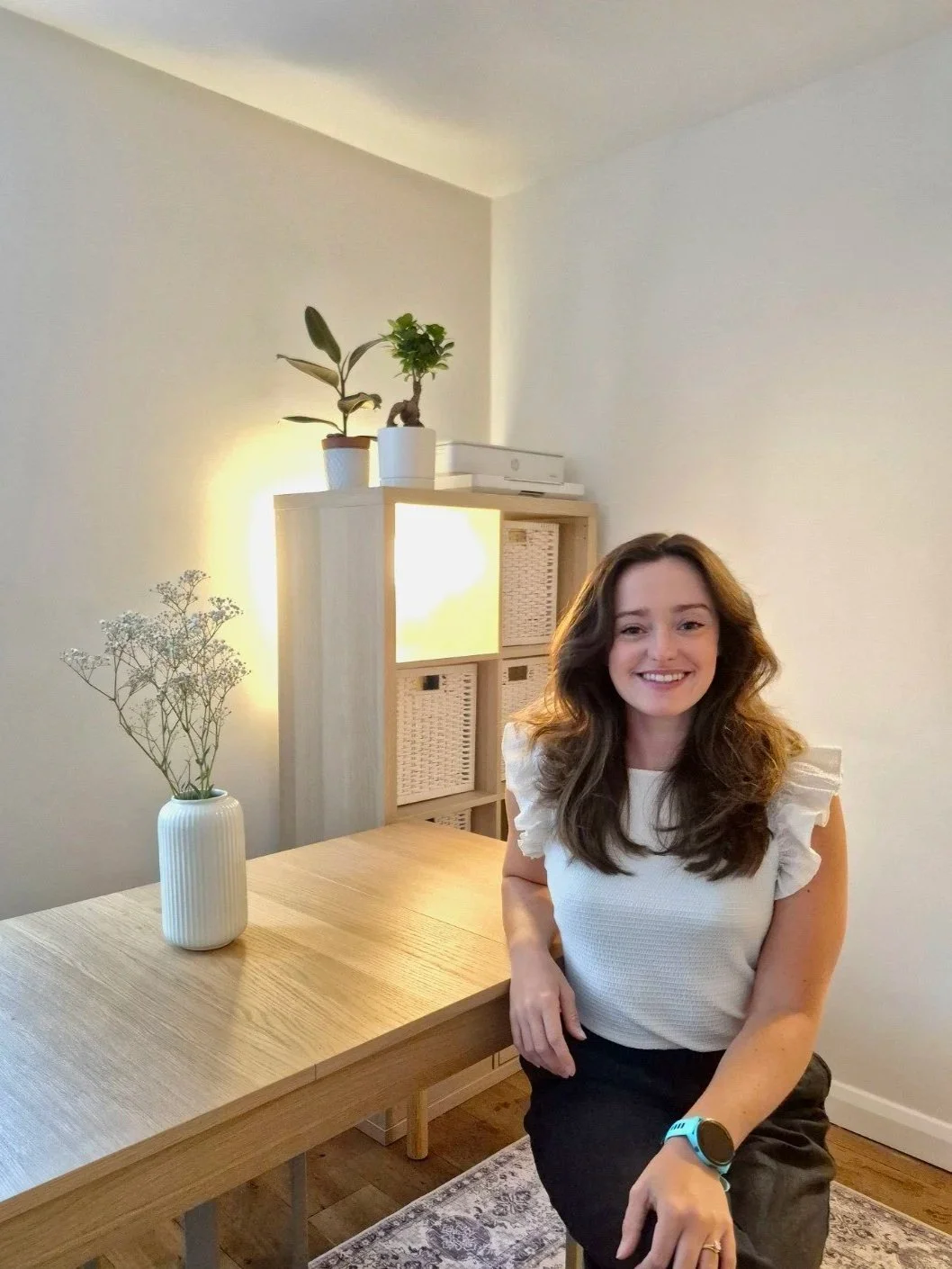A woman sitting at a wooden dining table with a white vase holding dried flowers in the foreground. In the background, there is a wooden shelf with three potted plants on top and indoor lighting illuminating the wall.