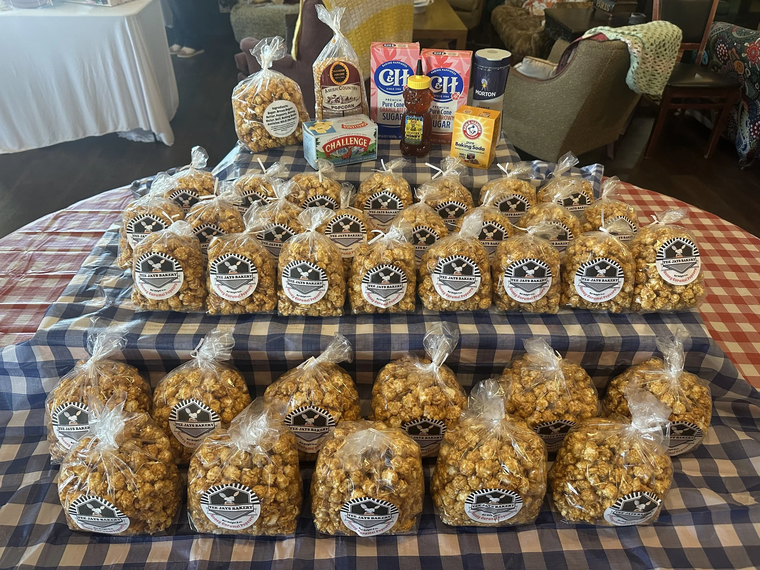 Assorted bags of caramel popcorn with a variety of snack items and baking ingredients on a checkered tablecloth in a cozy room.
