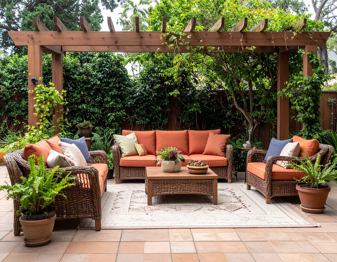 Outdoor patio with wicker furniture including a couch and chairs with orange cushions, a wooden coffee table with a floral centerpiece, and potted plants, under a wooden pergola surrounded by greenery.