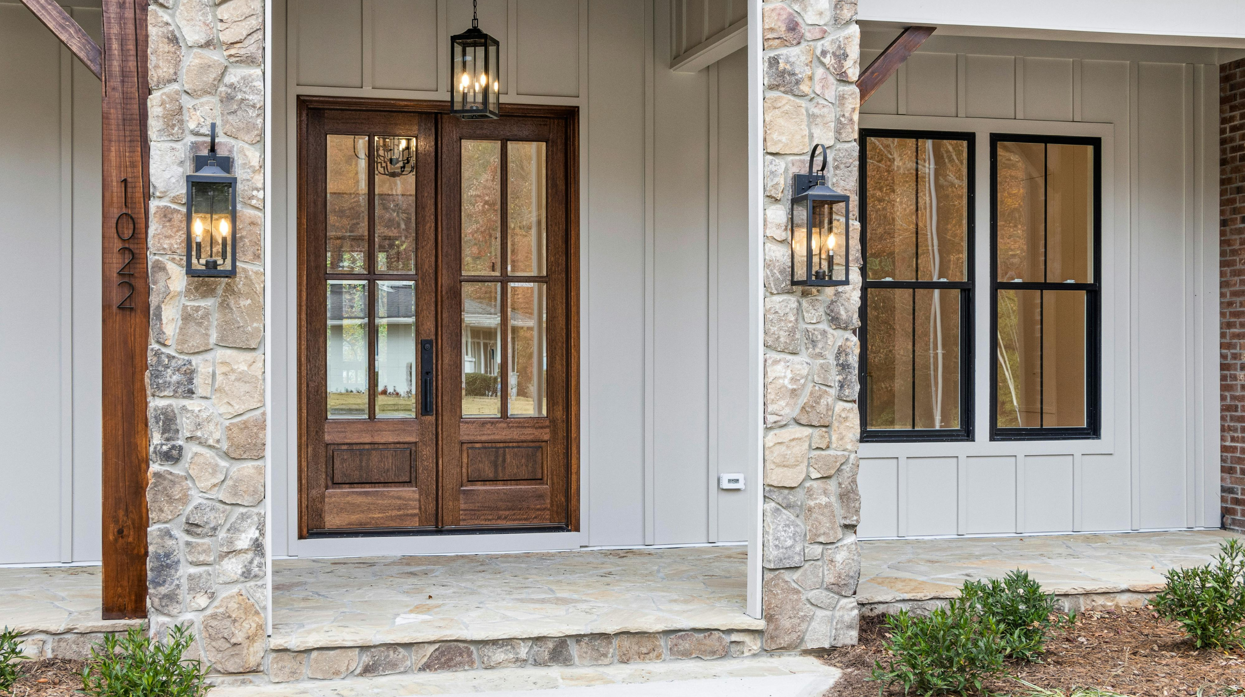 Front porch of a house with a wooden door with glass panels, stone columns, wall-mounted lanterns, and a window with black framing. The house number is 1022.