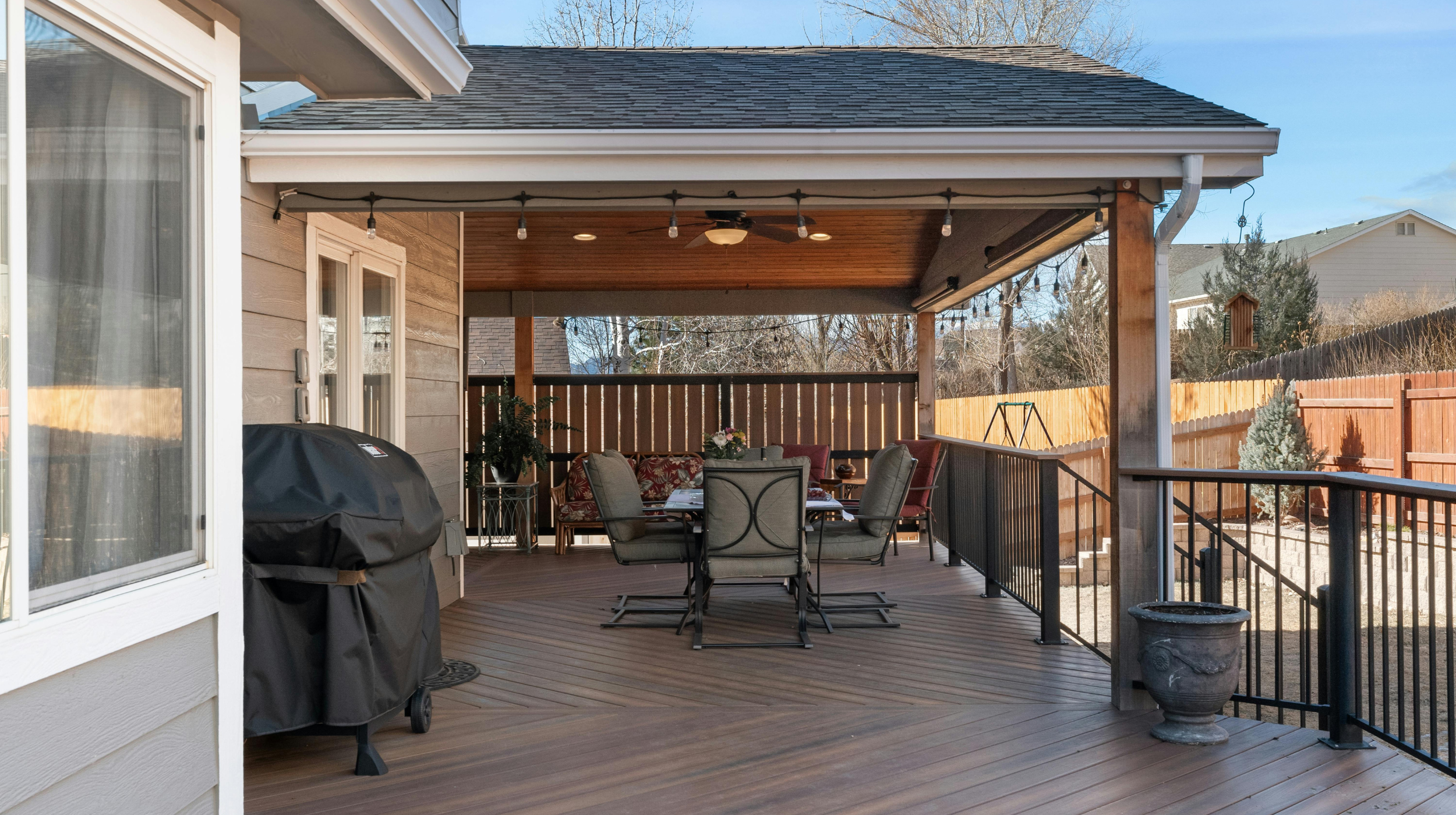 A covered outdoor patio area with a dining table, chairs, and string lights, attached to a house with beige siding and a black railing, with a neighboring house and trees in the background.