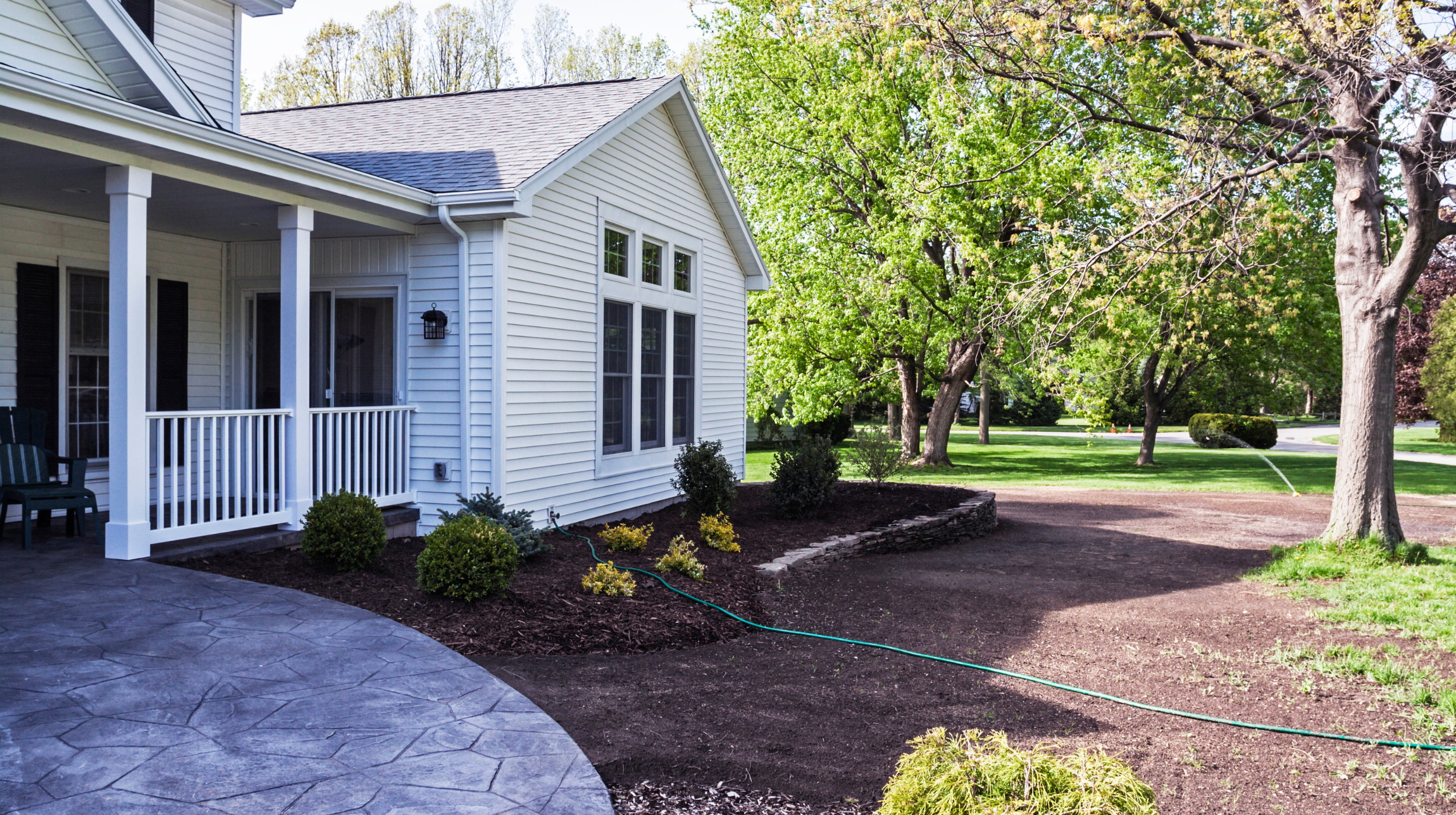 Backyard view of a white house with a porch, surrounded by green trees and a lawn, with some shrubs and a garden bed in the foreground.