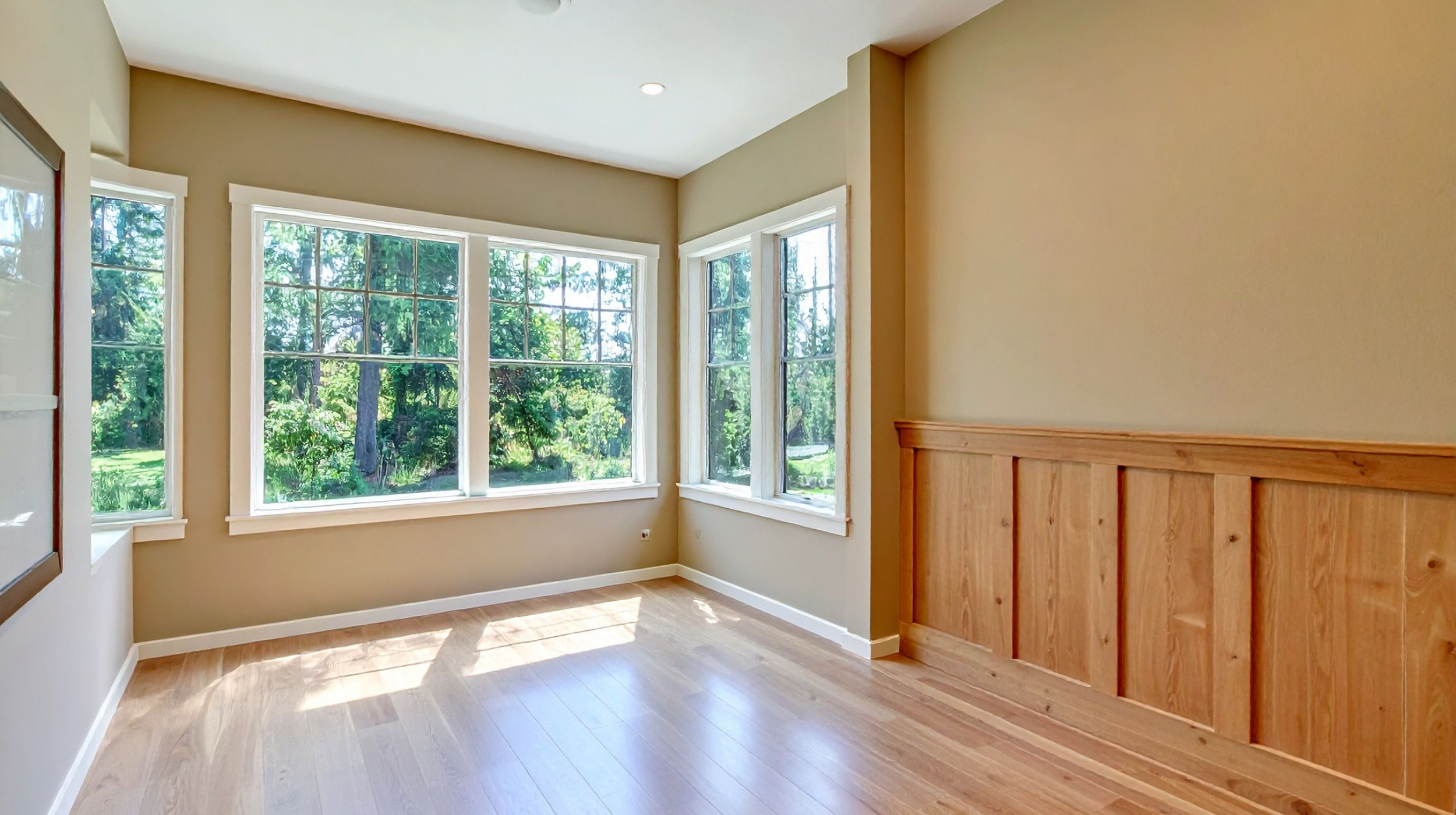 Empty room with large windows showing a view of green trees, beige walls, wooden wainscoting, and hardwood floors.