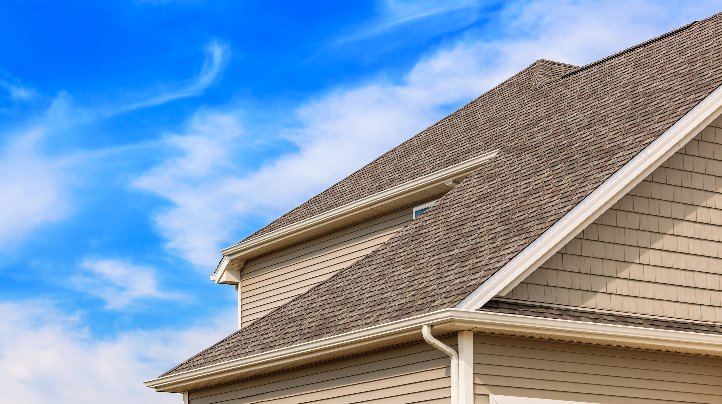 The image shows the side of a house with a beige exterior, vinyl siding, and a brown shingle roof against a bright blue sky with some clouds.