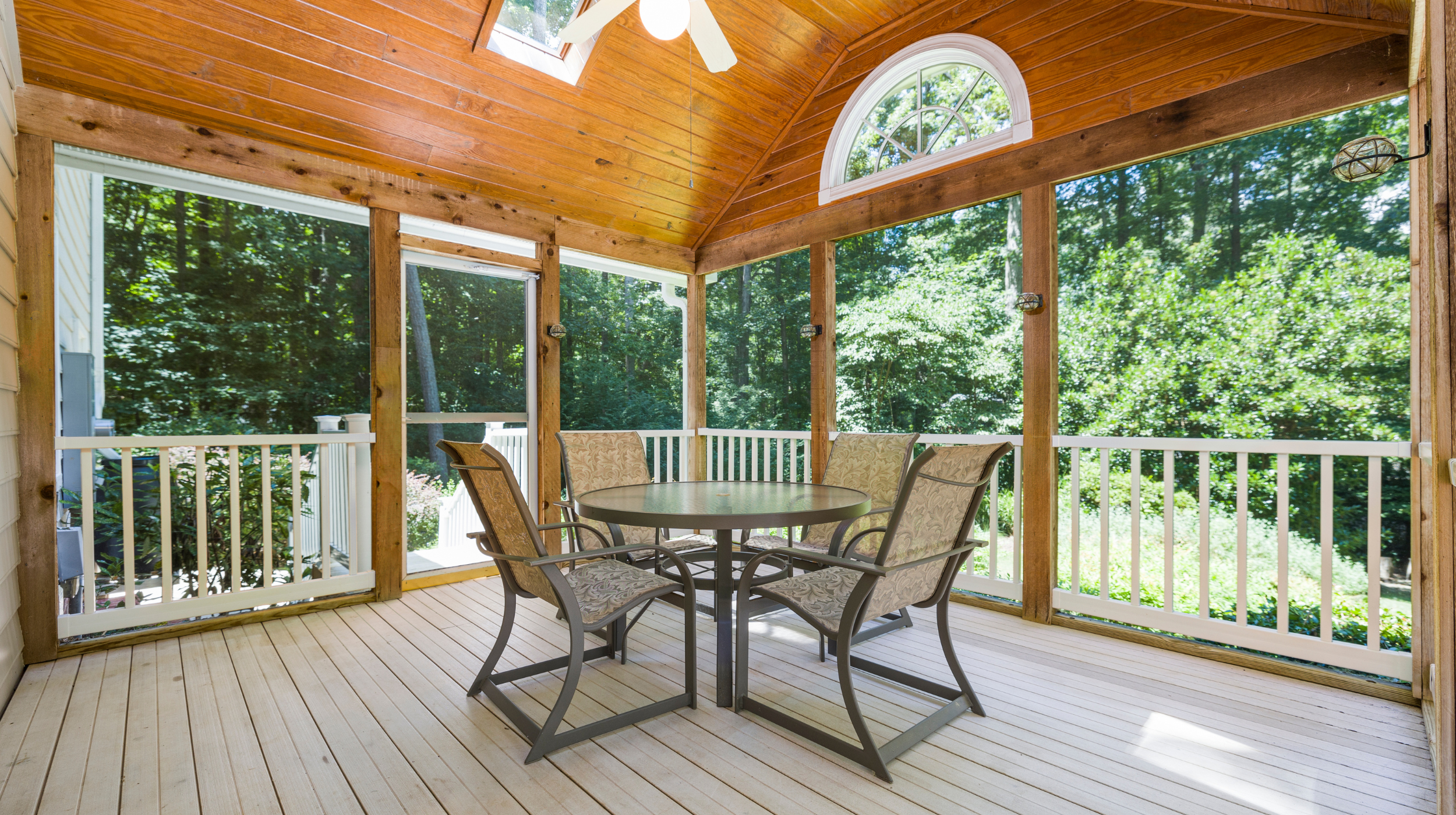 A screened porch with a round table, four chairs, wooden flooring, a skylight, and large screened windows overlooking green trees.