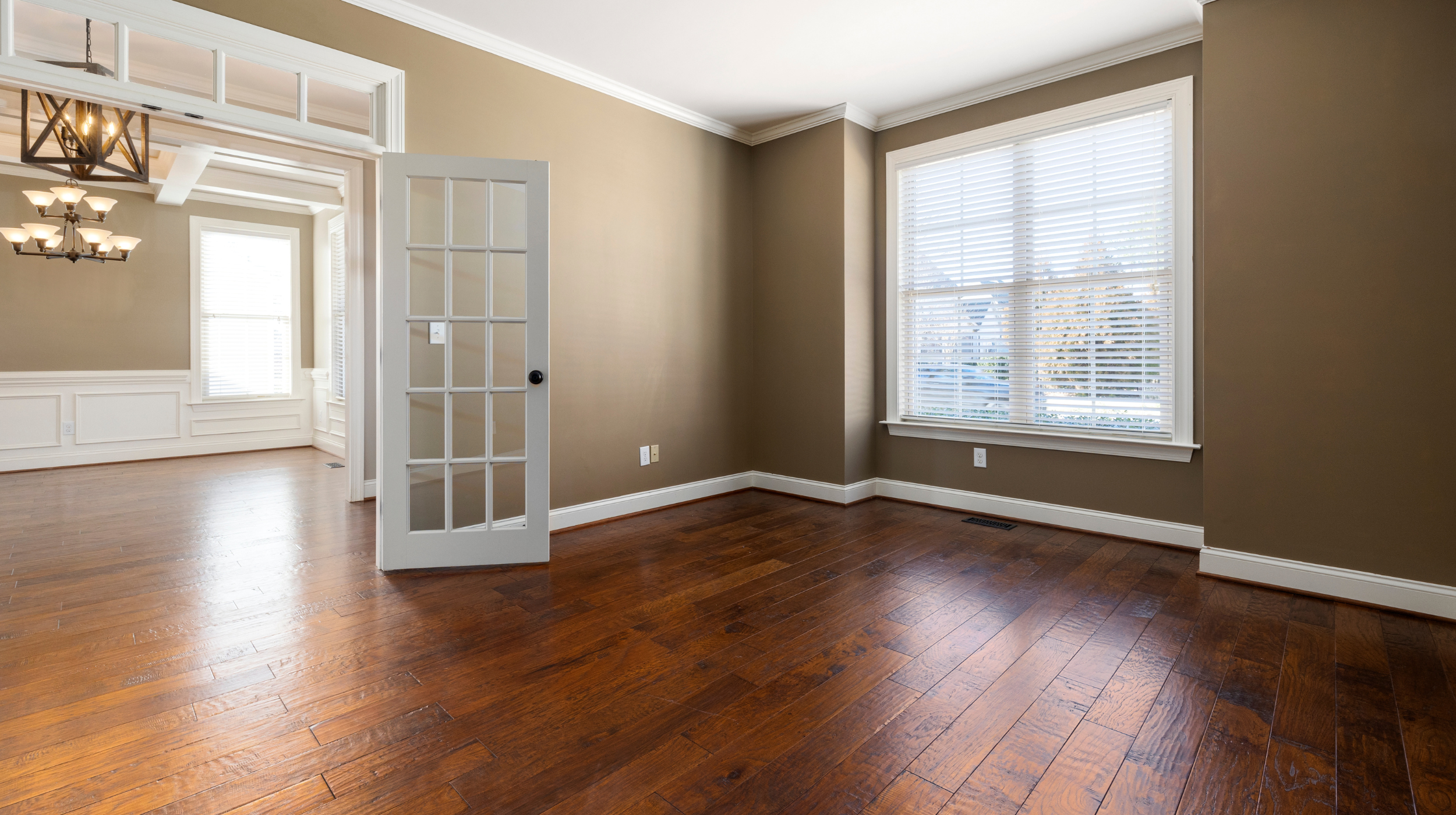 Empty living room with large windows, hardwood floors, beige walls, white trim, and a glass-paneled door.
