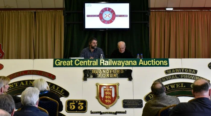 Two men sitting at a desk during a Railwayana auction, with a screen above displaying the auction logo, and signboards on the front of the desk reading 'Great Central Railwayana Auctions' and 'Blandford Forum.' Audience members are visible in the foreground.