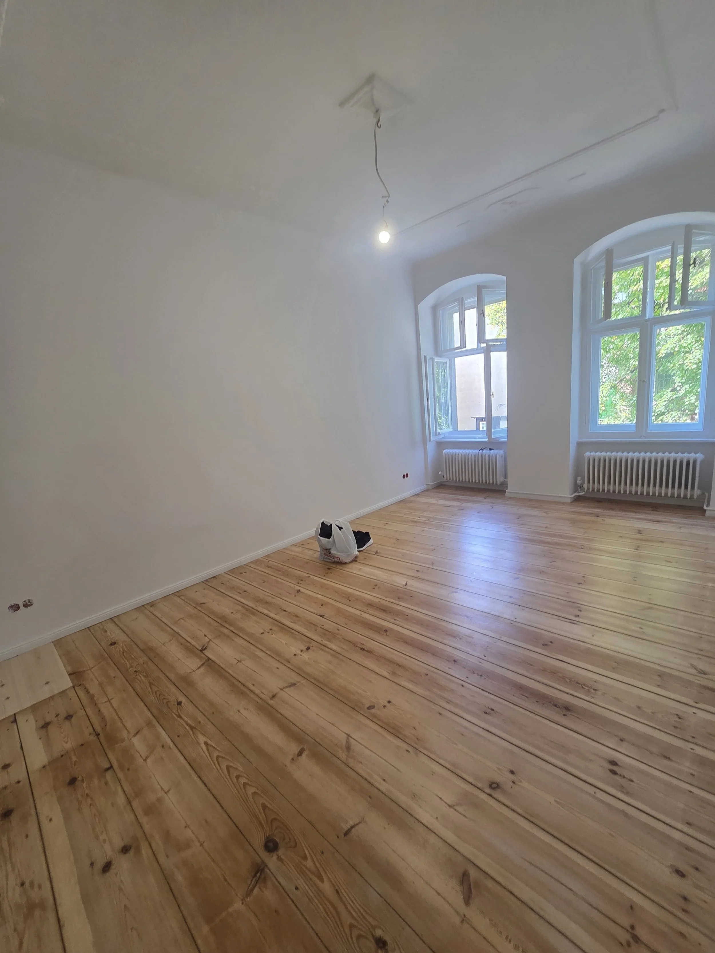 Room with unfinished walls, large windows, hardwood floors, and a single light bulb hanging from the ceiling.