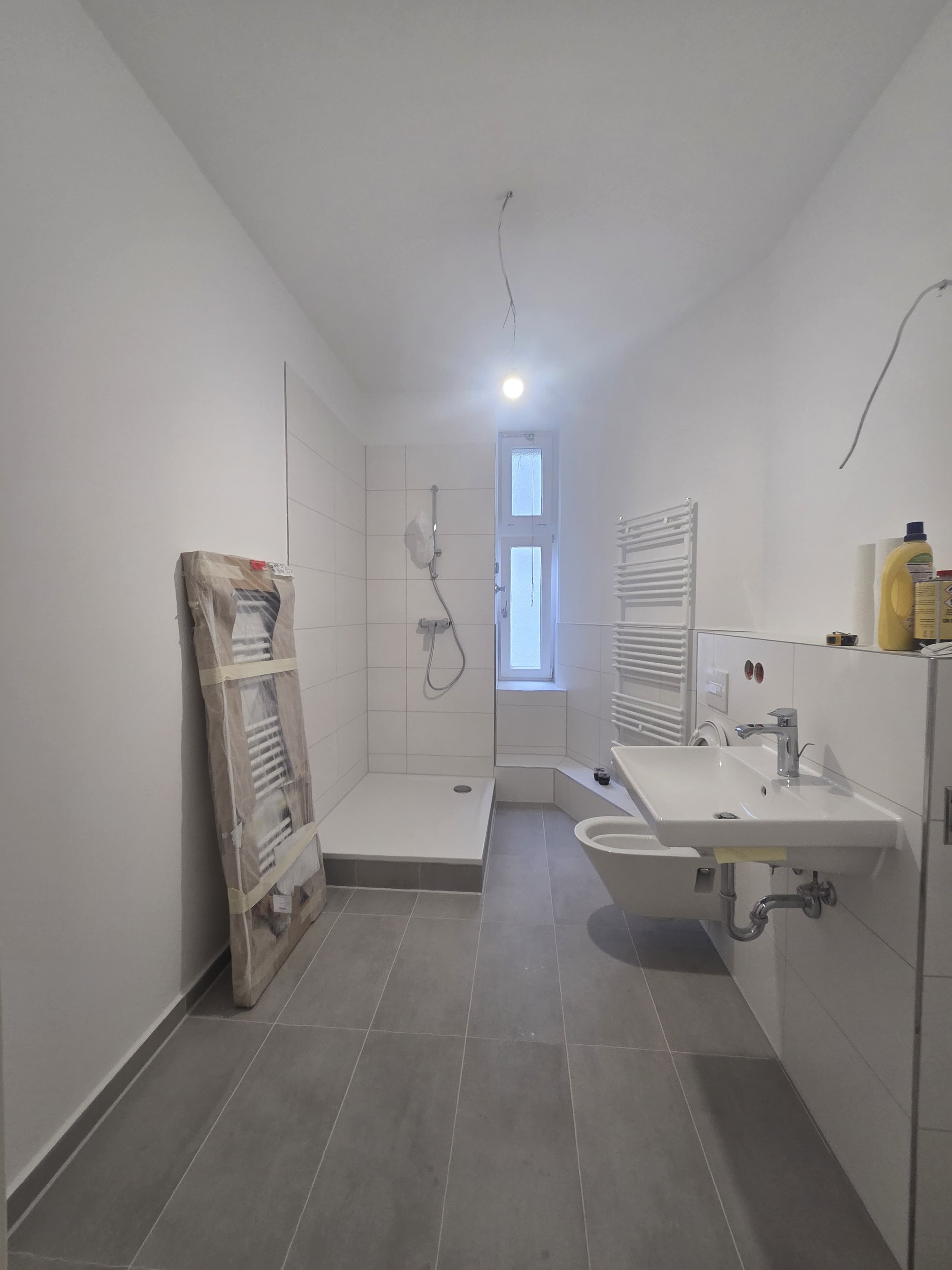 Bathroom under construction with gray floor tiles, white walls, a shower area, a white radiator, a window, a sink, and various toiletries and construction items.