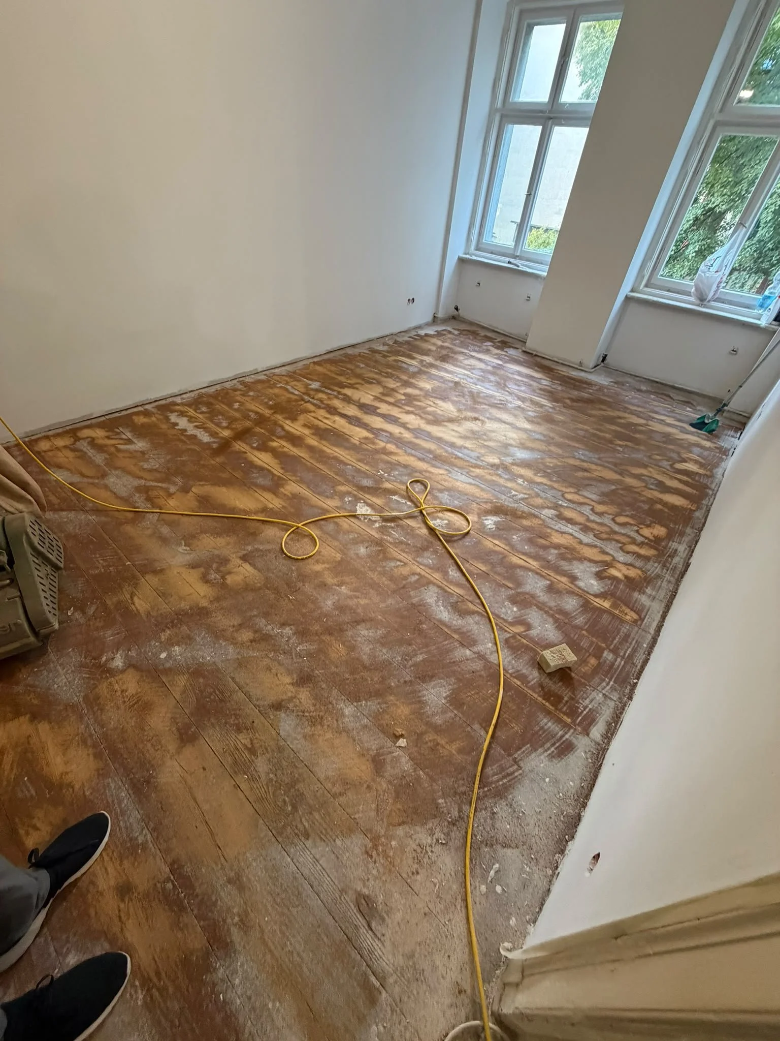 Room with hardwood floors being sanded down, a yellow extension cord on the floor, and large windows with trees visible outside.