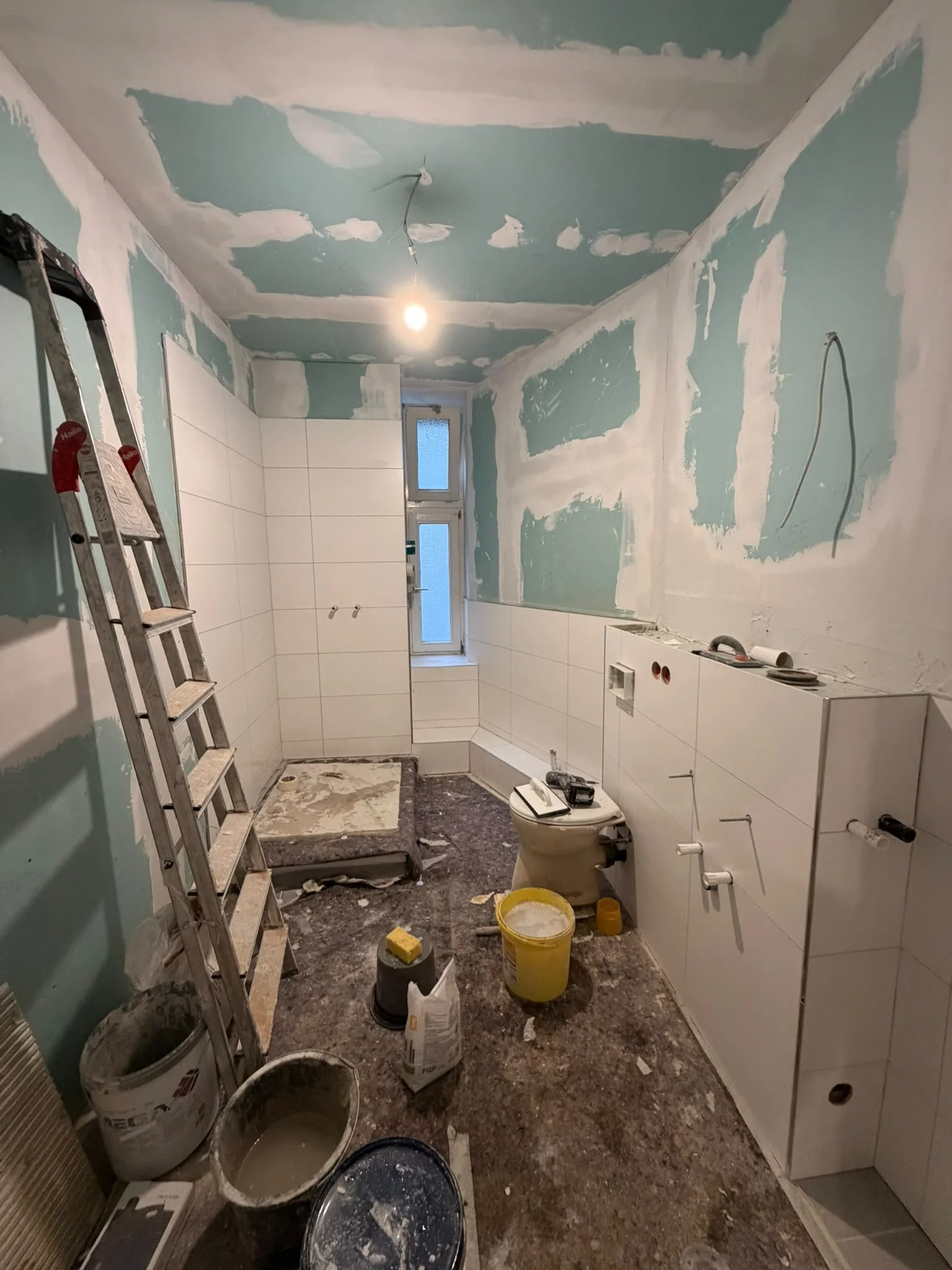 A bathroom under renovation with unfinished drywall, construction tools, and supplies scattered on the floor, and a small window at the far end.