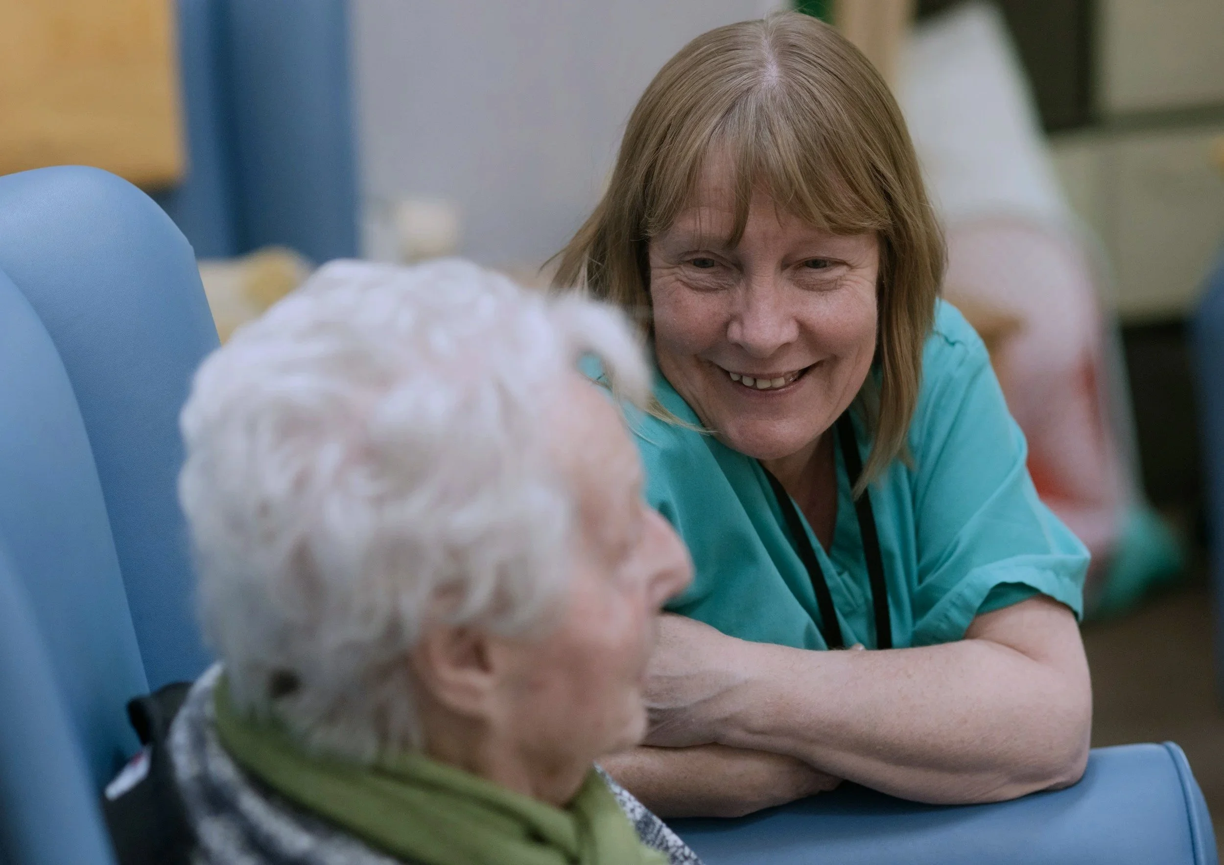 A woman with light brown hair smiling and leaning over to talk to or interact with an elderly person with white hair, both sitting at a table in a room that appears to be a care facility or hospital.