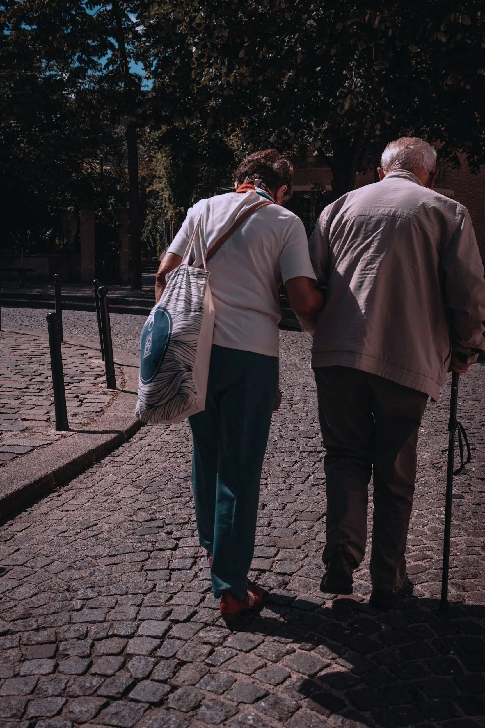 Two elderly people, a woman and a man, walk together on a cobblestone street in a shaded area with large trees in the background. The woman carries a tote bag with a swirl pattern, and both are dressed casually.