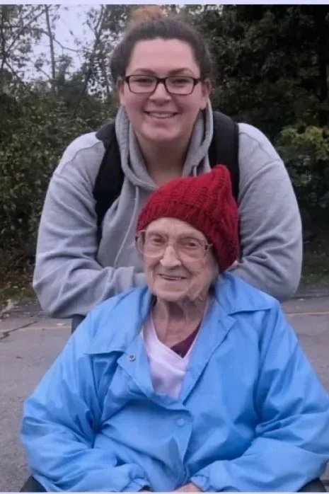 A young woman with glasses and a gray hoodie smiling behind an elderly woman with glasses, wearing a red knit hat and a blue raincoat, seated outdoors.