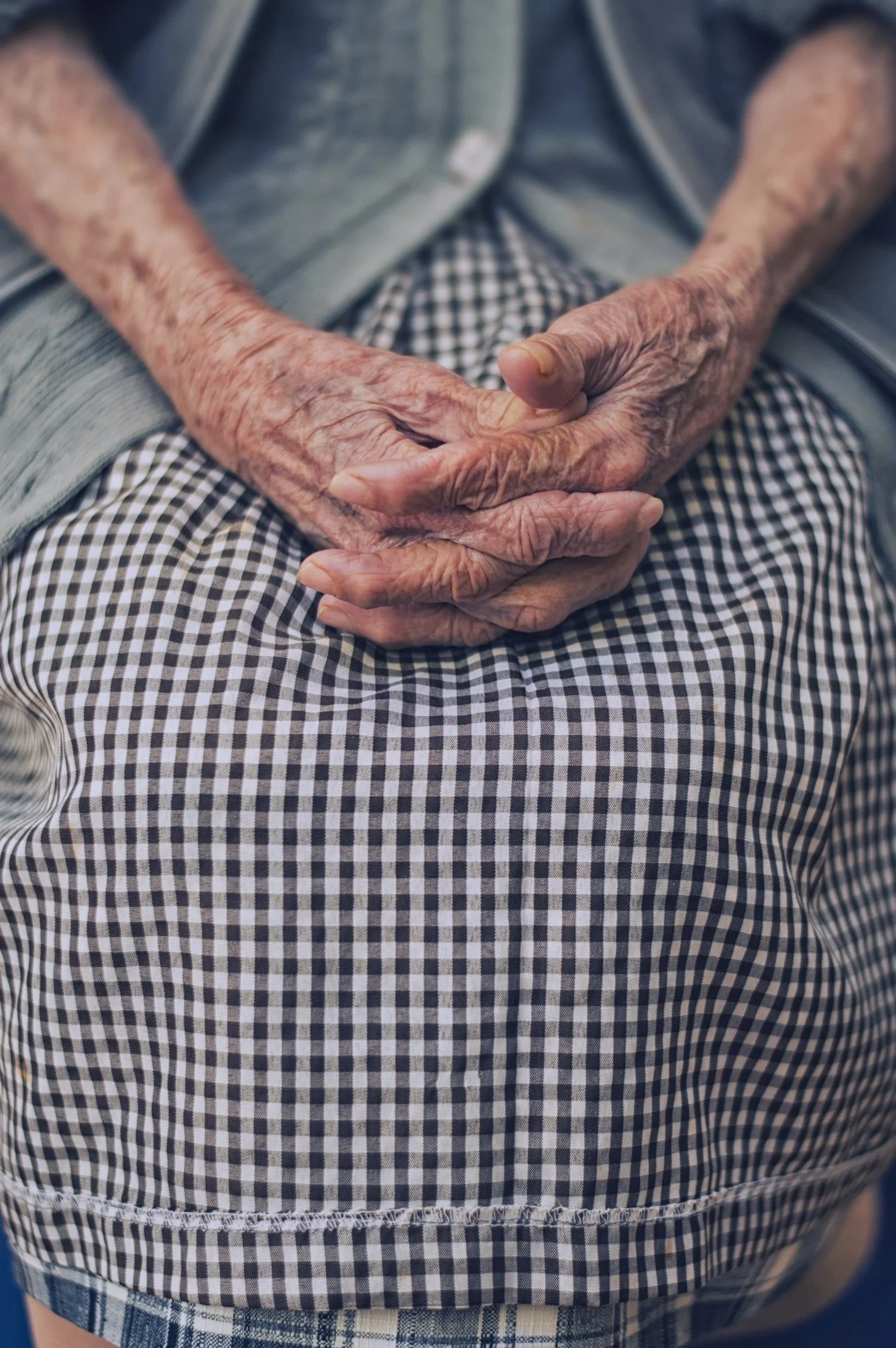 Close-up of elderly person's hands resting on their lap, wearing a checkered shirt and jeans.