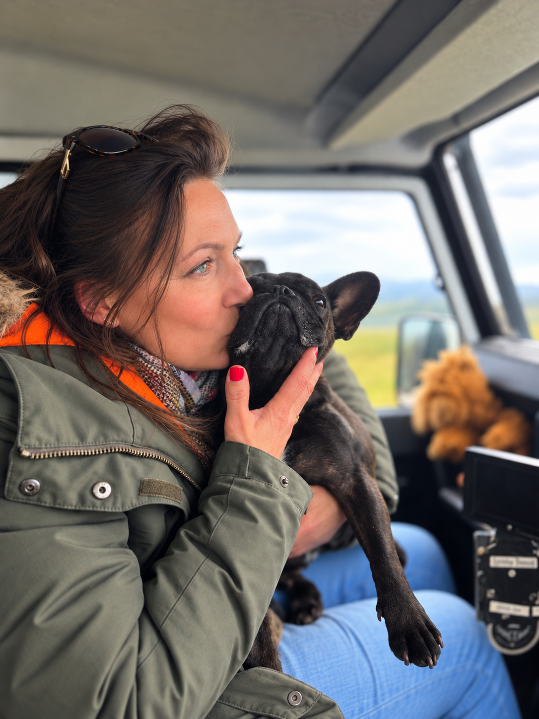 A woman holding a French Bulldog inside a vehicle, kissing the dog on the nose.