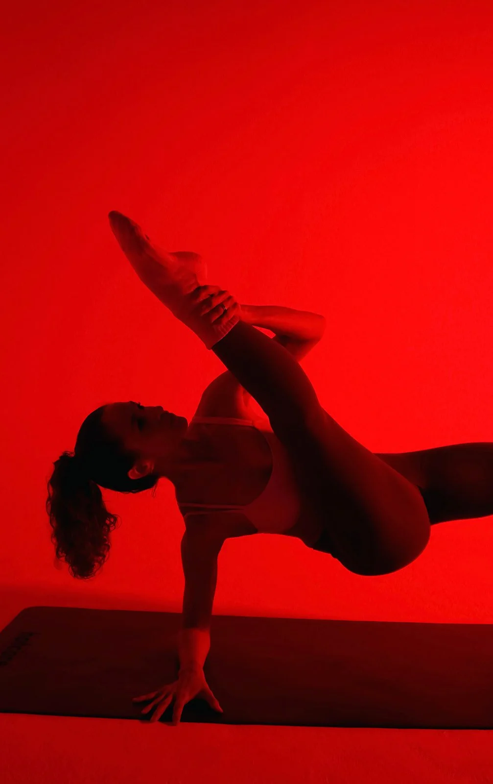 A woman in athletic wear performing a yoga pose on a yoga mat under red lighting.