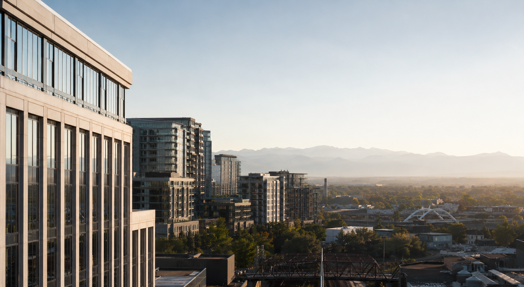 Cityscape with modern high-rise buildings, a bridge, and distant mountains under a clear sky.