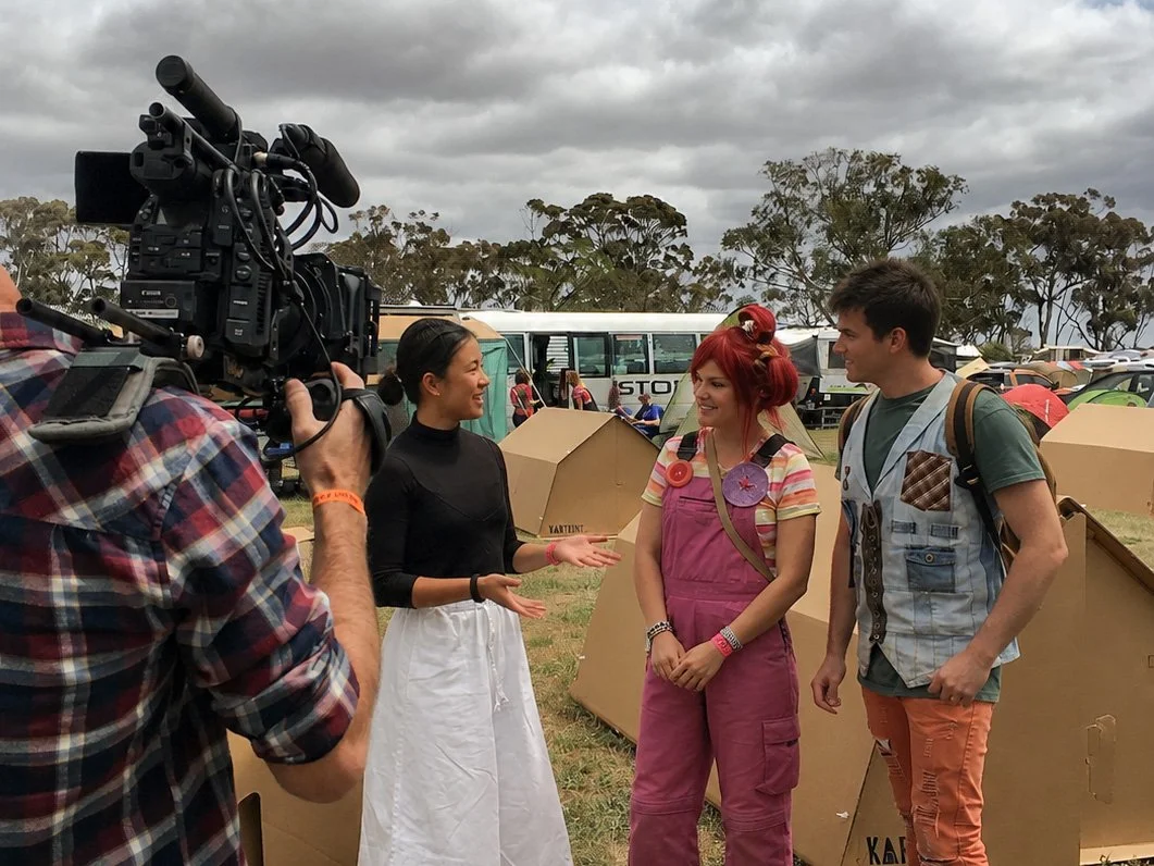 A cameraman filming three people talking outdoors at a festival or event on a cloudy day. The woman in the middle has red hair, wearing pink overalls and striped shirt, and is smiling. The woman on the left has dark hair, wearing a black top and white skirt. The man on the right has dark hair, wearing a short-sleeved shirt with patches, and orange pants.