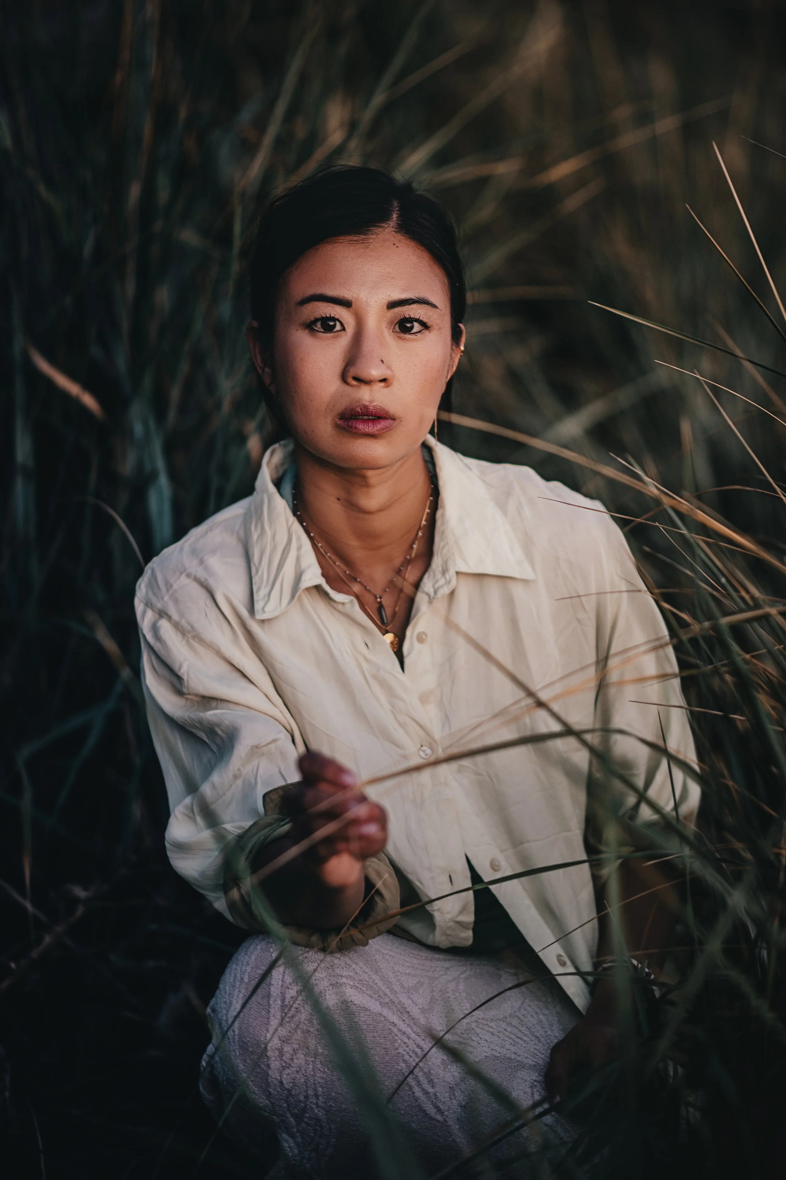 A woman with dark hair, wearing a light-colored shirt and layered necklaces, is sitting outdoors among tall grass or plants at dusk, looking directly at the camera with a neutral expression.