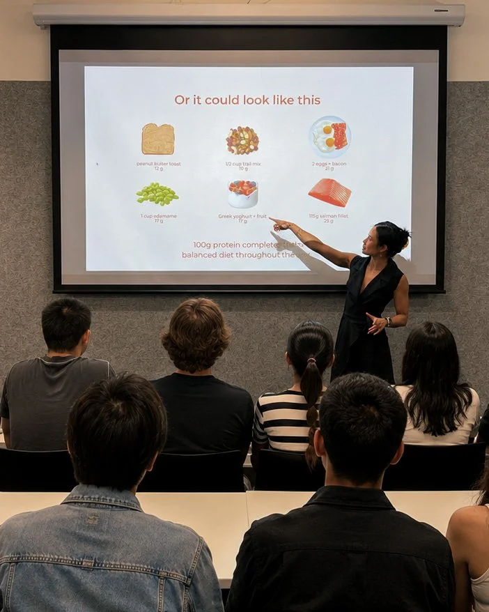 A woman giving a presentation in a classroom with a slide showing nutritious foods and portion sizes. The slide includes images of peanut butter toast, trail mix, bacon and eggs, edamame, Greek yogurt with fruit, and salmon fillet.