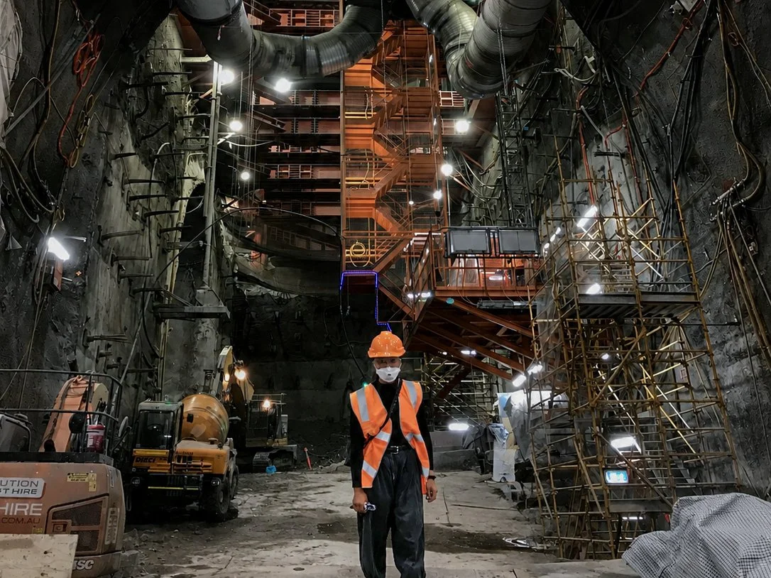 Construction worker in safety gear and face mask standing inside an underground tunnel with scaffolding, pipes, and construction equipment.