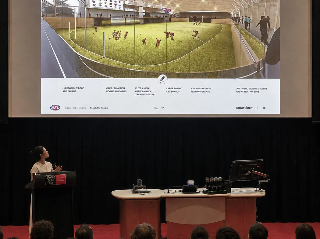 A woman is giving a presentation on a stage with a large screen showing a design plan for an indoor sports facility. The plan includes a field, seating, and other amenities. The audience is seated in front of the stage.