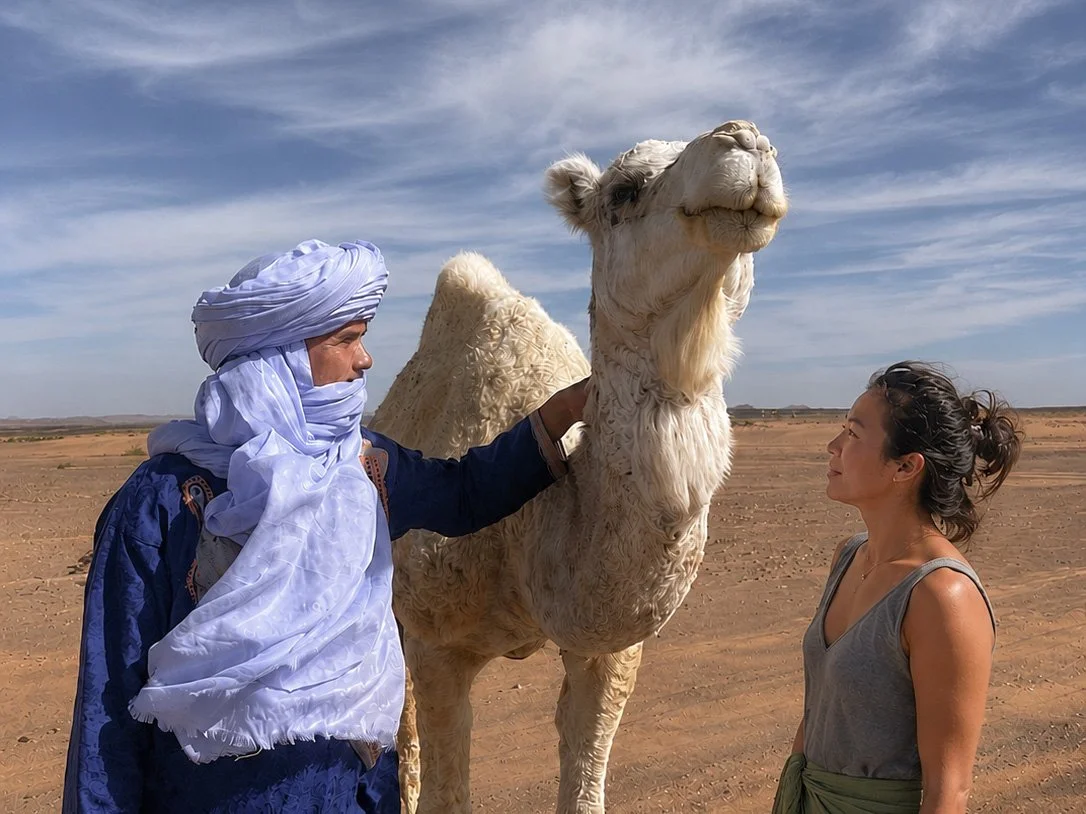 A man in traditional desert attire and a woman face a camel in a desert landscape, with a blue sky and scattered clouds.