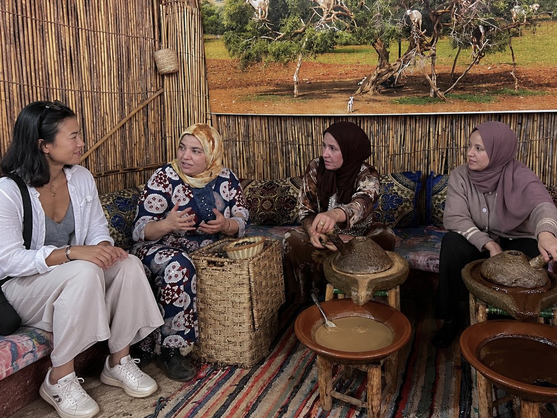 Four women sitting and talking inside a traditional yurt with woven walls, with pottery and baskets around them, and a landscape with trees visible through an opening in the wall.