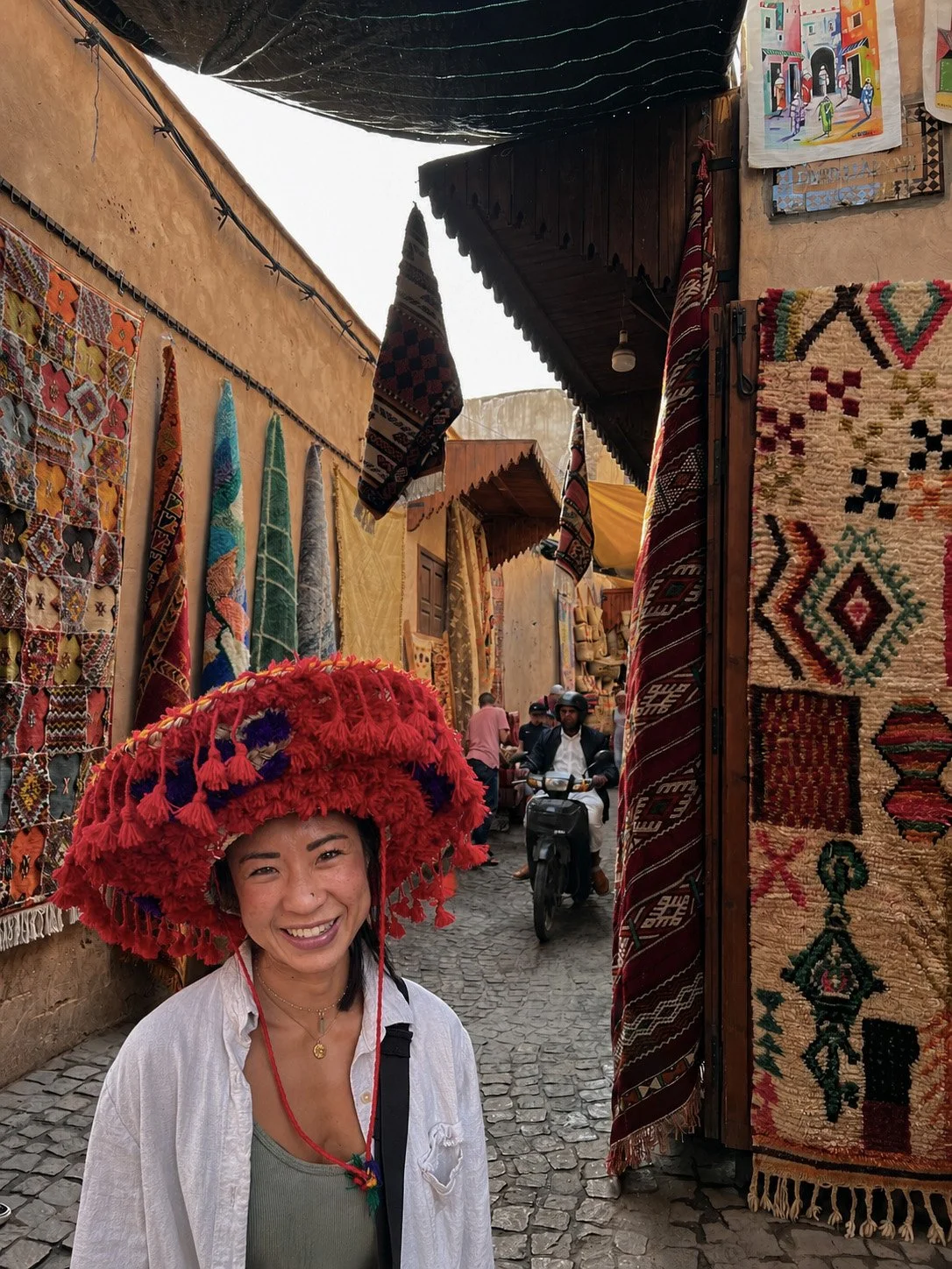 A woman smiling and wearing a large red, tasseled hat in a marketplace with colorful rugs and textiles hanging on the walls, and people riding motorcycles in the background.