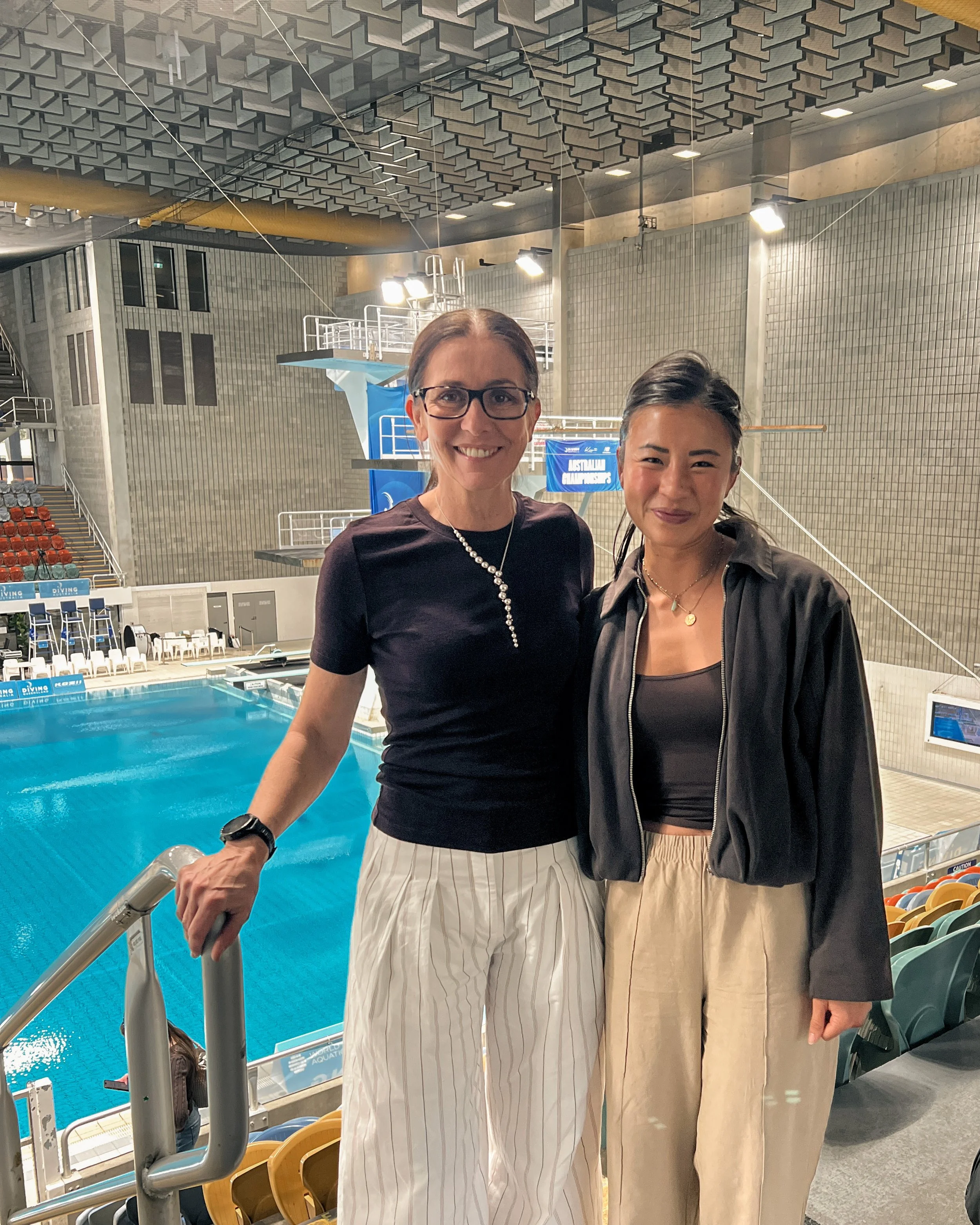 Two women smiling at an indoor swimming pool. The woman on the left wears glasses and striped pants, the one on the right wears beige pants and a black jacket.