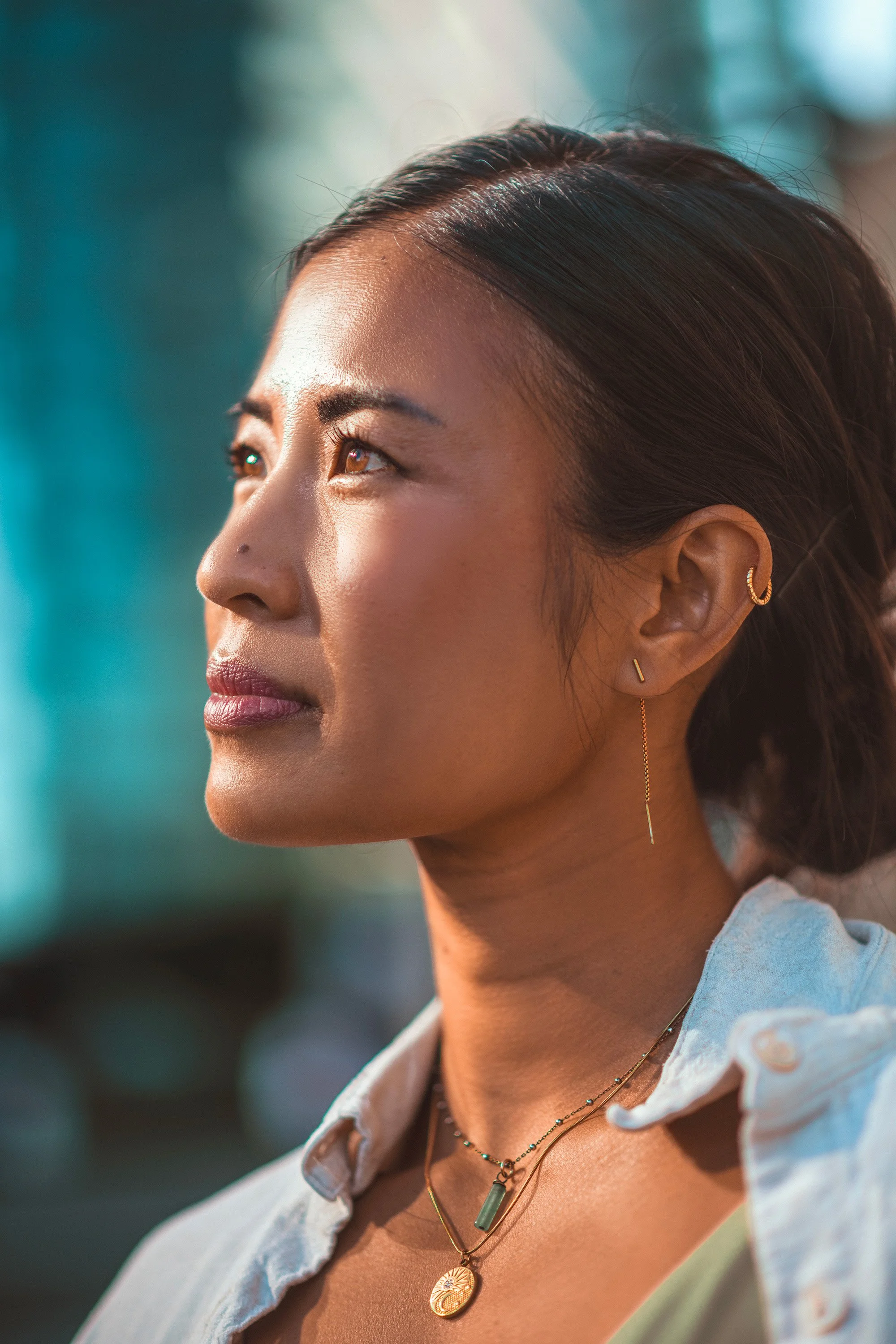 A close-up of a woman with dark hair, wearing gold earrings and layered necklaces, looking upward with sunlight on her face.