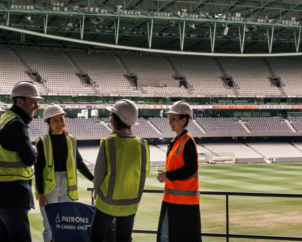 Four construction workers, three women and one man, wearing safety helmets and reflective vests, standing and talking inside a large empty stadium with a grass field.