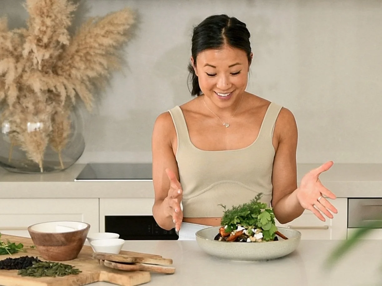 A woman in a beige tank top is smiling and presenting a colorful salad in a bowl in a modern kitchen.