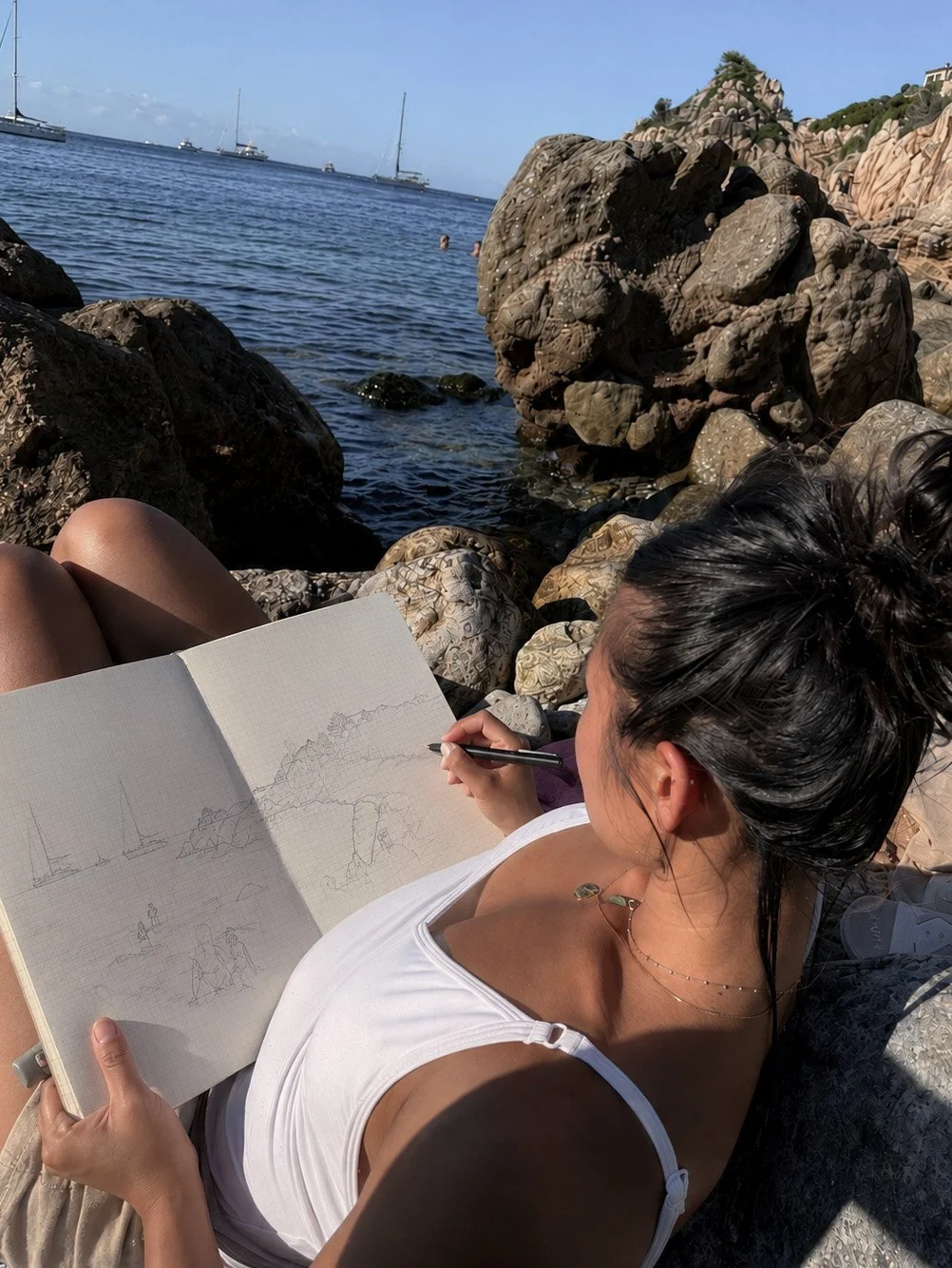 A woman sitting on rocks at the beach, sketching in a notebook, with large rocks and the ocean in the background, and sailboats anchored in the water.