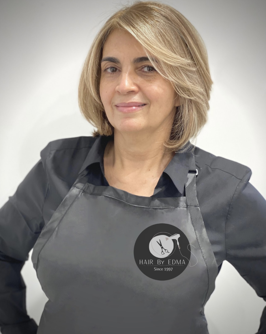 A woman with blonde hair wearing a black shirt and a dark apron with the logo of a hair salon, 'HAIR By EDMA Since 1997', smiling at the camera against a plain white background.