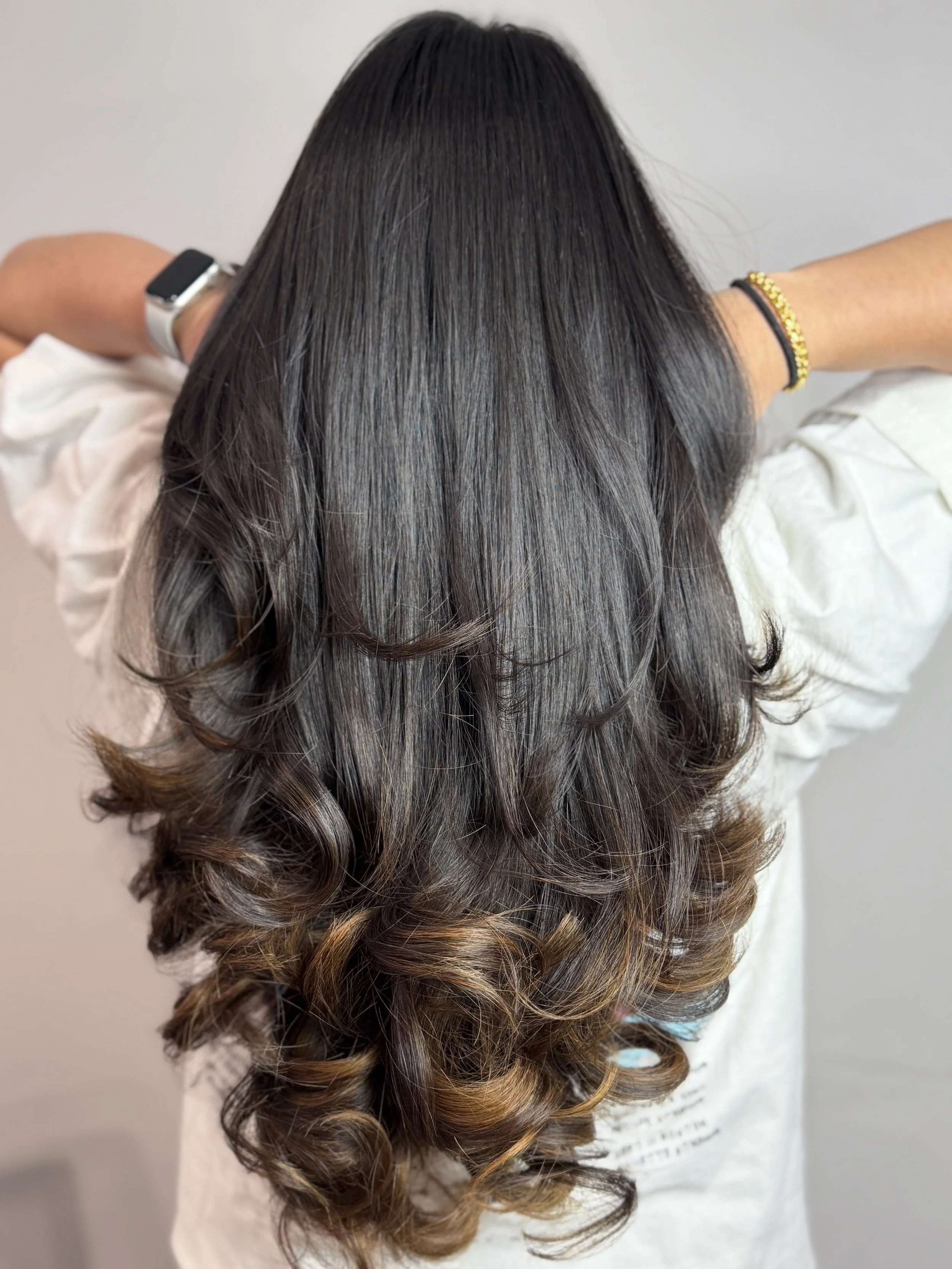 A woman with dark brown hair that is straight on the top and styled in loose curls at the ends, wearing a white top, with a black and gold bracelet on her wrist, adjusts her hair with her arms raised behind her head against a plain light-colored back
