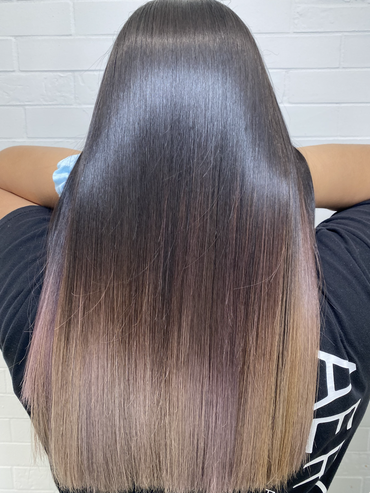Person with long, straight, shiny hair that is dark at the top and gradually transitions to light brown or blonde at the ends, sitting against a white brick wall.