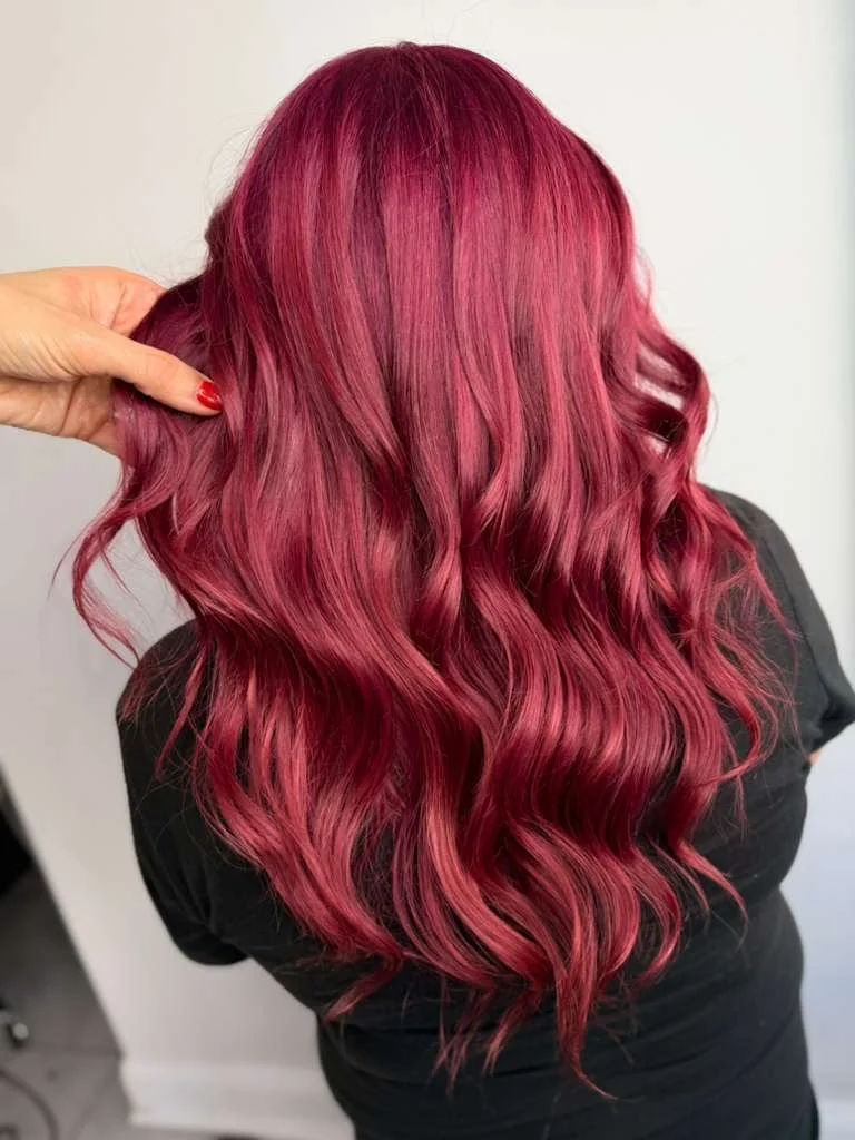 Back view of a woman with long, wavy, pinkish-red hair, holding a strand with her hand.