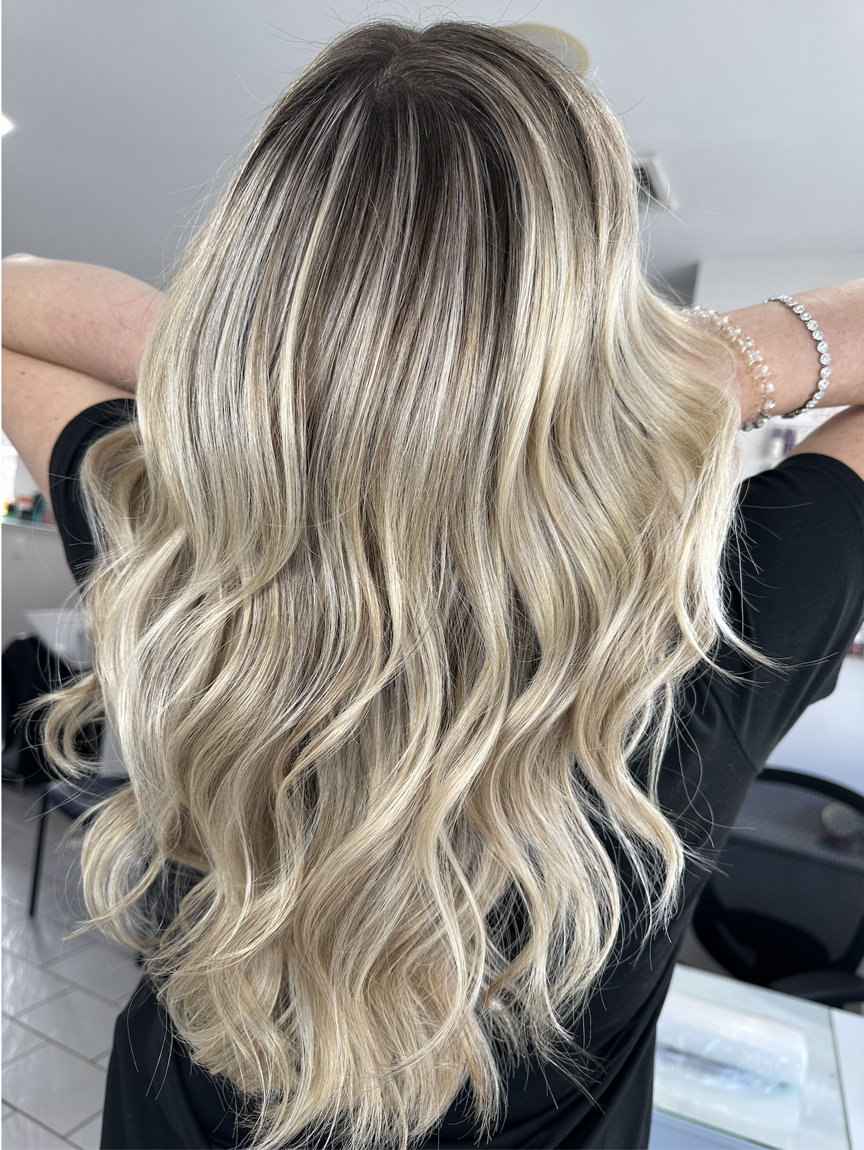 Woman with long, wavy blonde hair with darker roots, wearing a black top and a pearl bracelet, in a salon setting.
