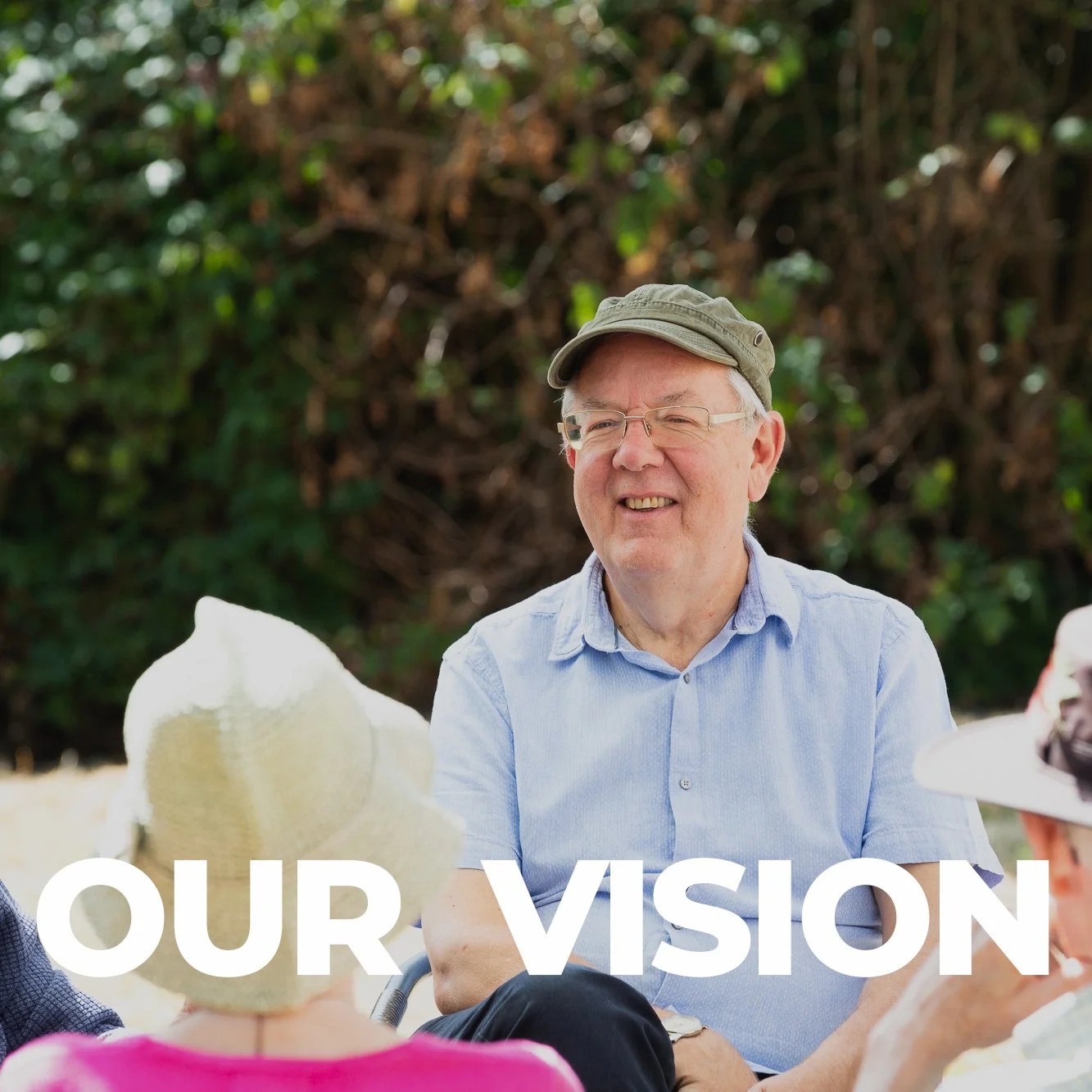 A man with glasses and a green cap, smiling, sitting outdoors with a group of people, with text that says 'OUR VISION' over the bottom part of the image.