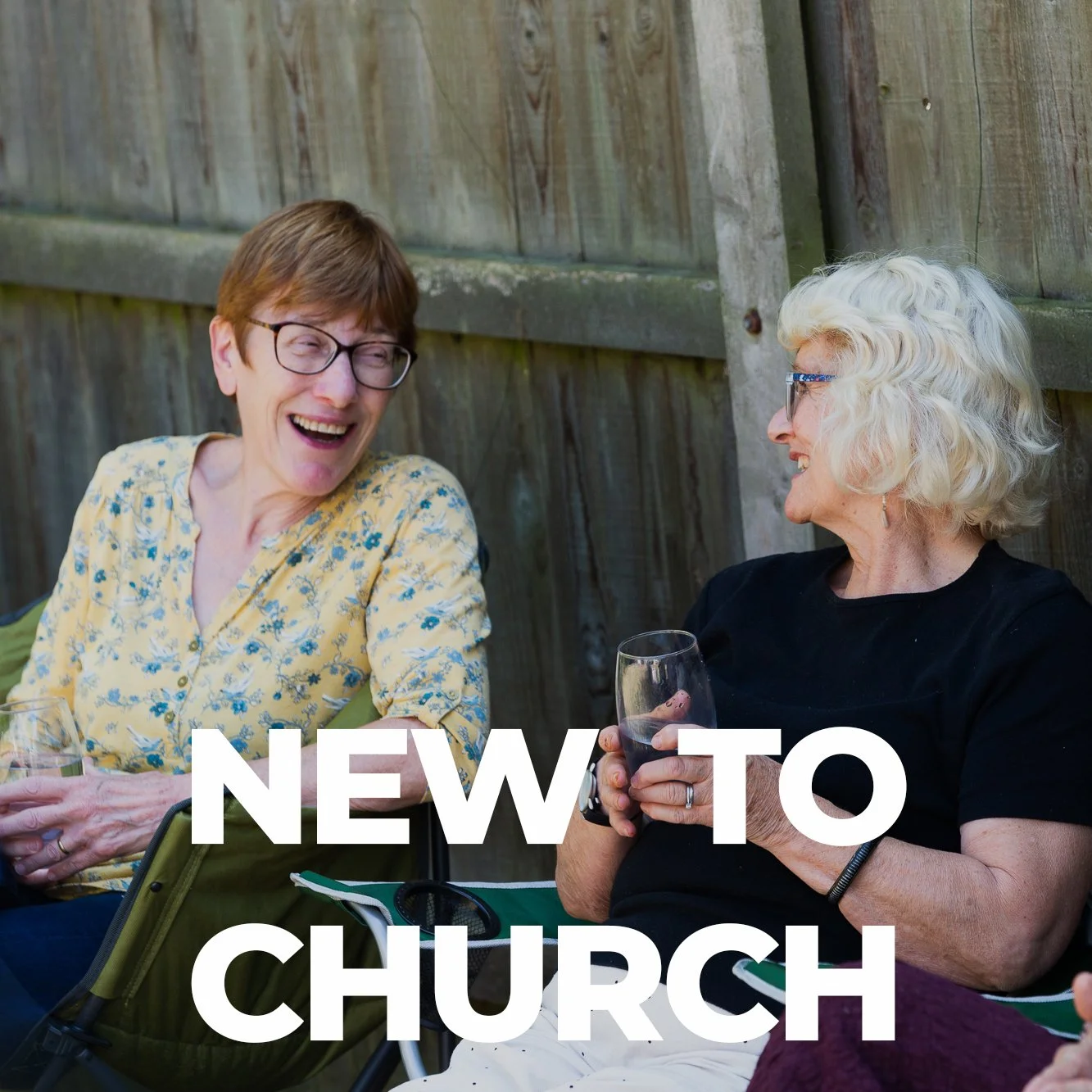 Two women sitting outdoors, laughing and talking, one holding a glass of red wine, with a wooden fence in the background.
