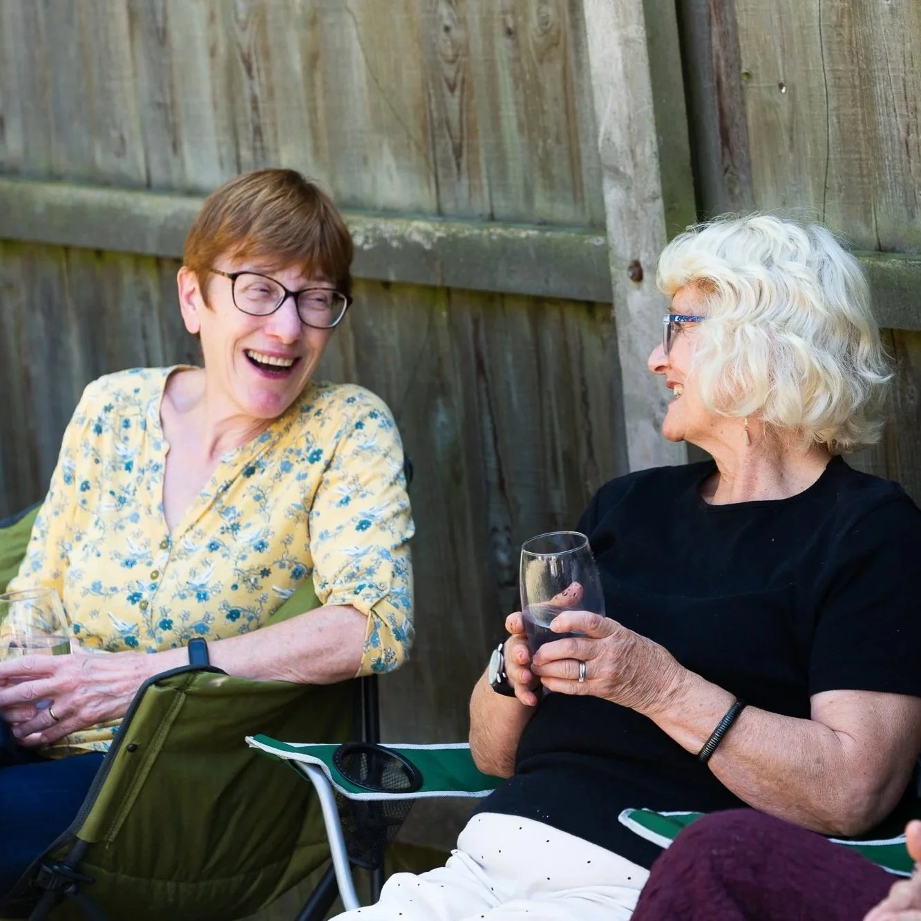 Two elderly women sitting outdoors against a wooden fence, smiling and talking. One woman has short brown hair, glasses, and a yellow floral shirt, the other woman has curly white hair, glasses, and a black shirt, holding a glass of red wine.