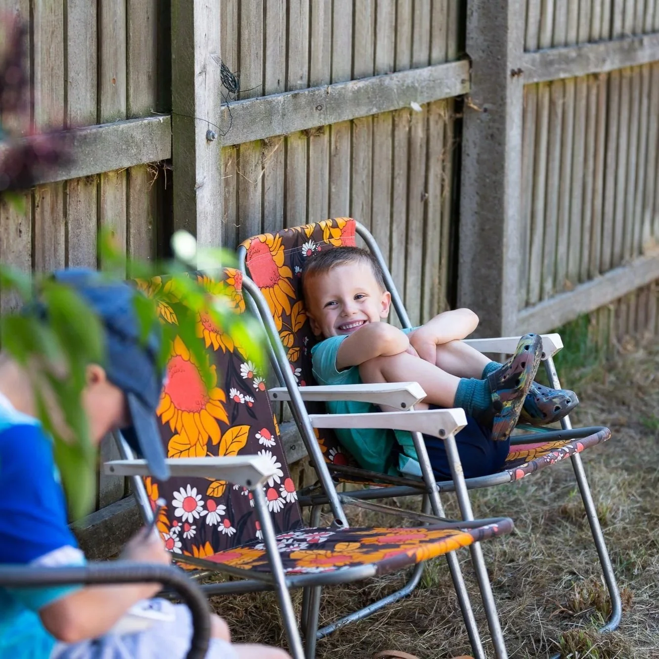 A young boy smiling and sitting in a garden chair with a floral pattern, leaning back and holding his knees, outdoors near a wooden fence, with another person partially visible in the foreground.