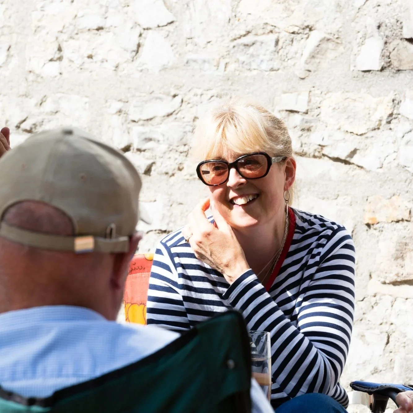 A smiling woman with blonde hair wearing large sunglasses and a striped shirt, talking to an older man with a beige cap, against a stone wall background.